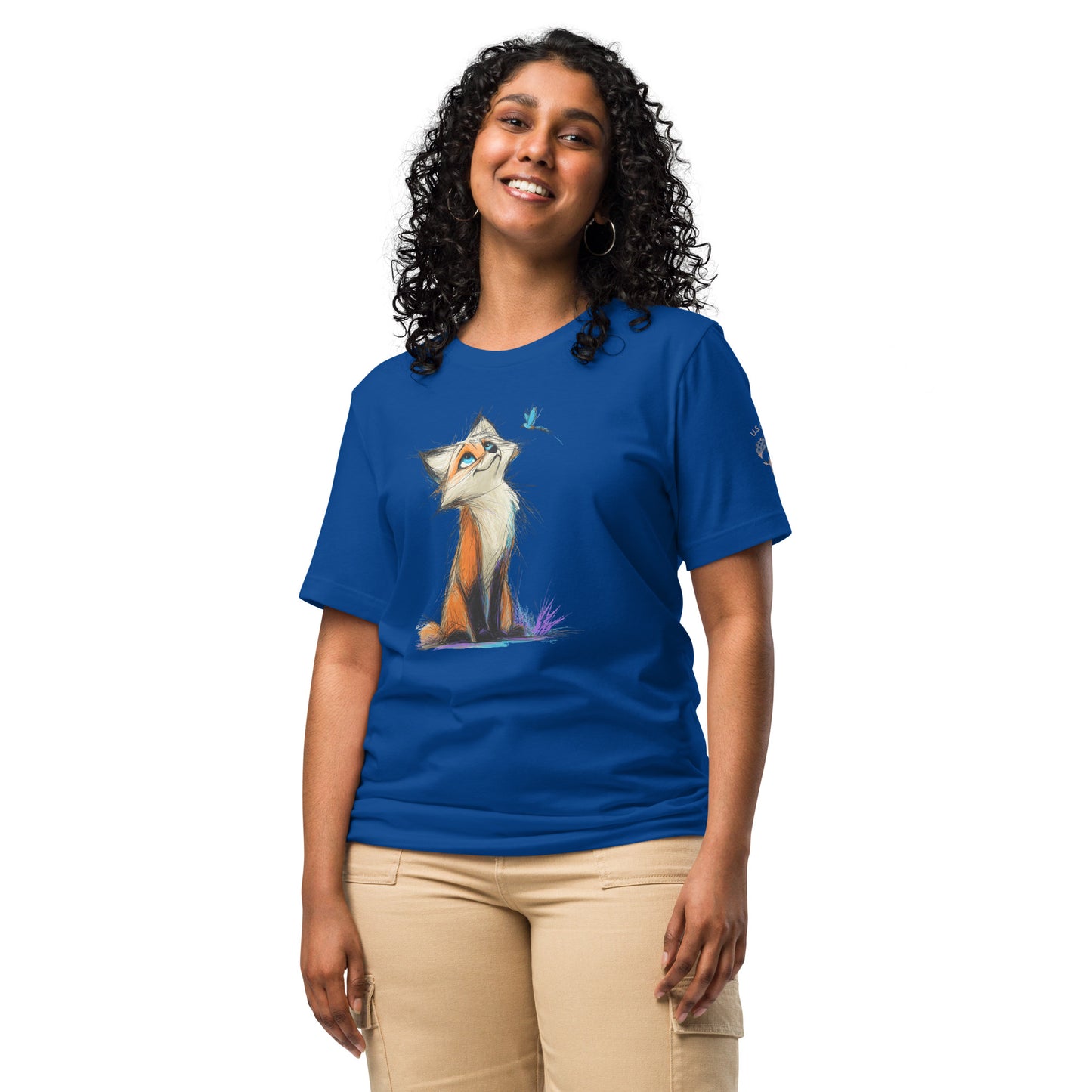 Product mockup. A woman in beige pants wearing a royal blue T-shirt with a whimsical orange-and-white cartoon fox looking up at a blue bird, set in blue and purple grass.