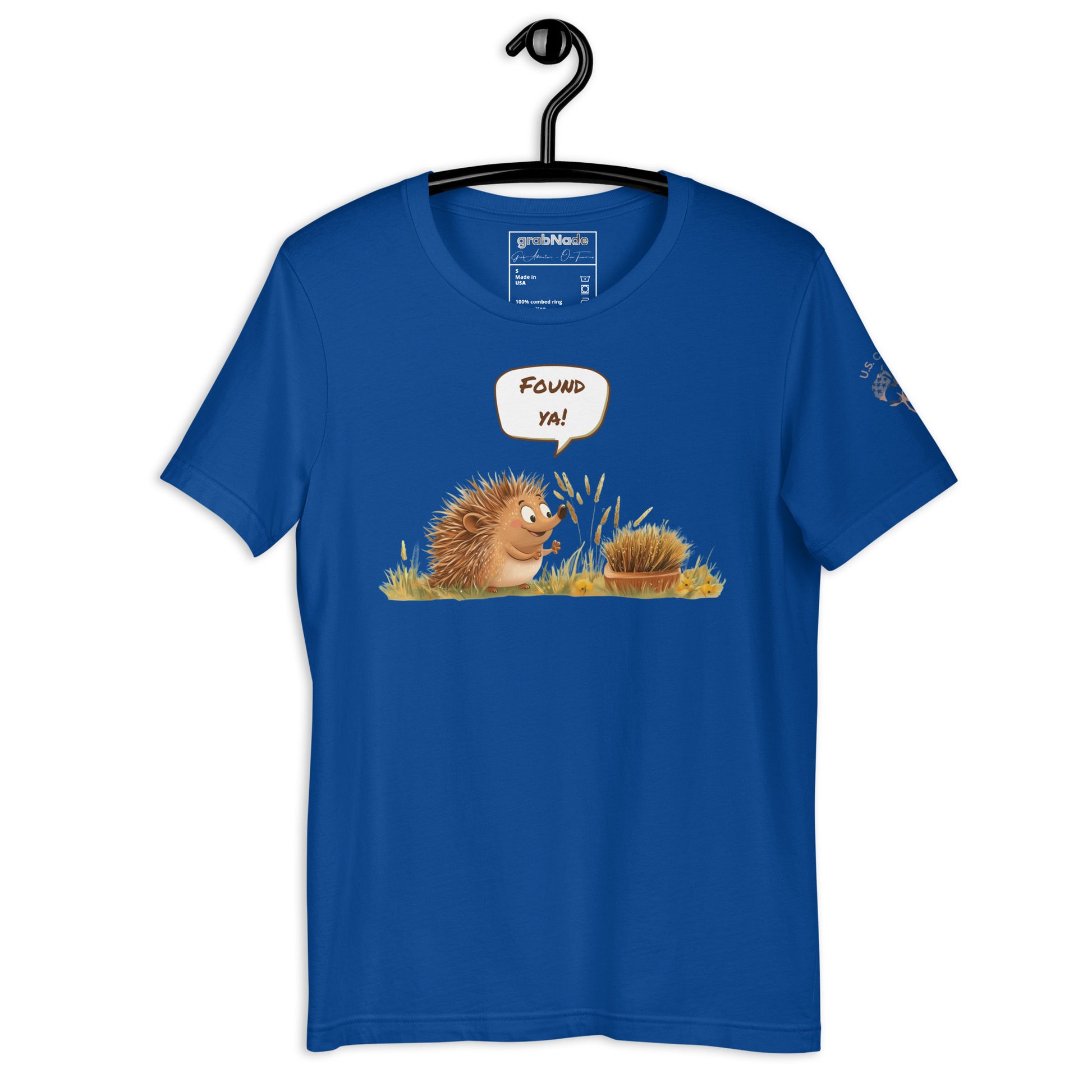 Product mockup. Blue T-shirt on a hanger showing twin cartoon hedgehogs, one says “Found ya!” above them, surrounded by yellow grass.