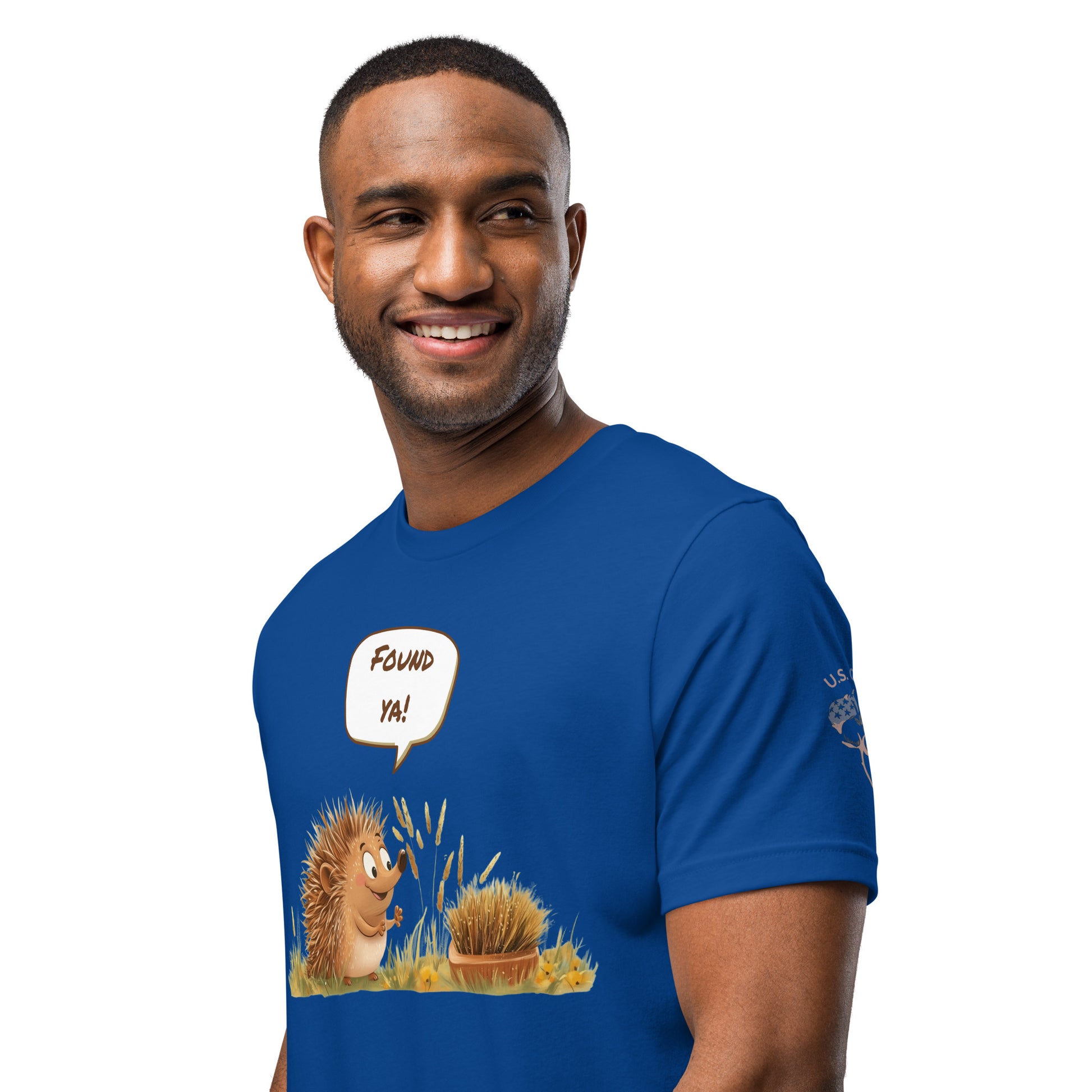 Product mockup.Blue T-shirt, left front, cartoon hedgehog saying “FOUND YA!” next to a disguised spiky ball in the grass, close-up from the torso up.
