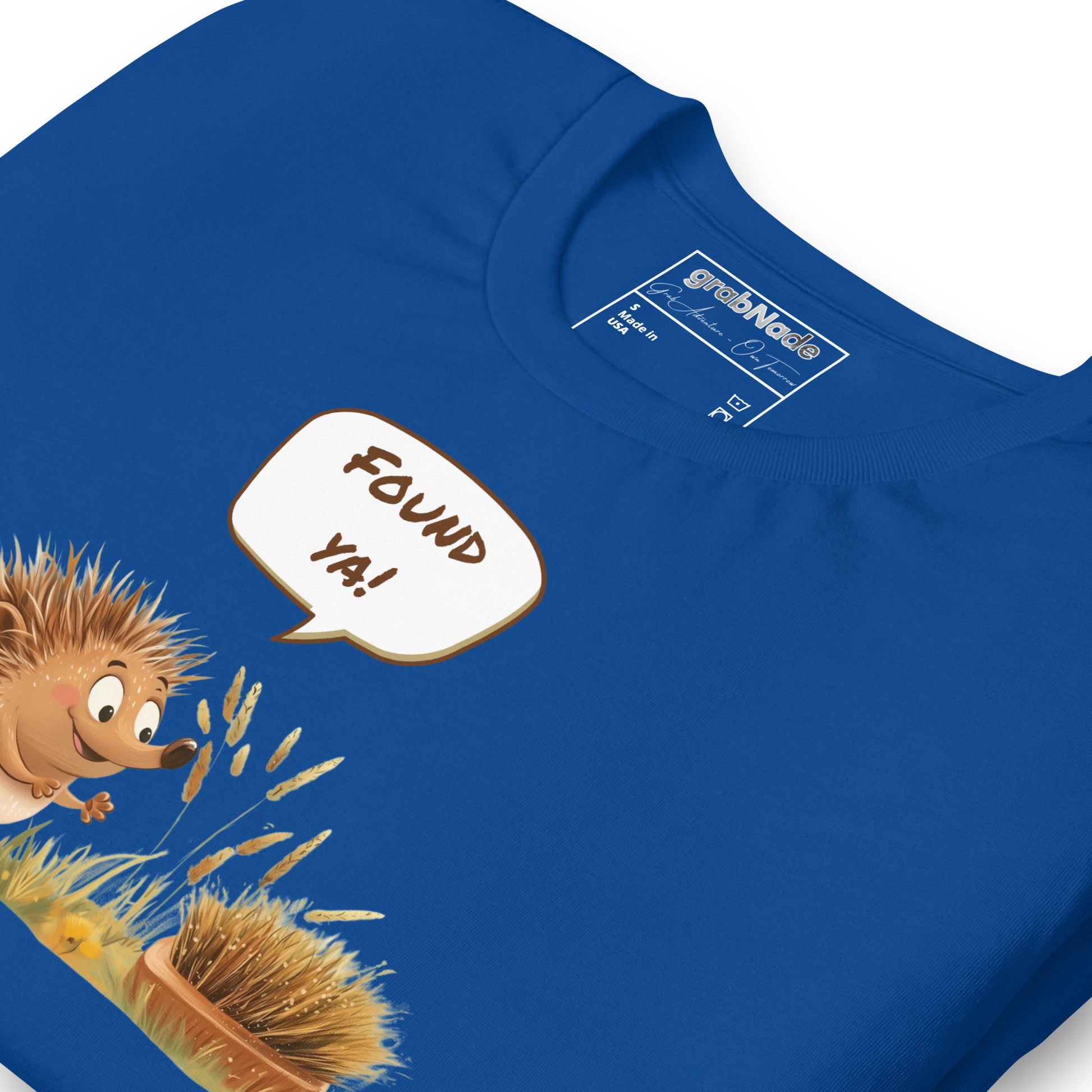 Product mockup. Blue T-shirt, zoomed in, cartoon hedgehog says “FOUND YA!” to a camouflaged brush in tall, yellow grass.