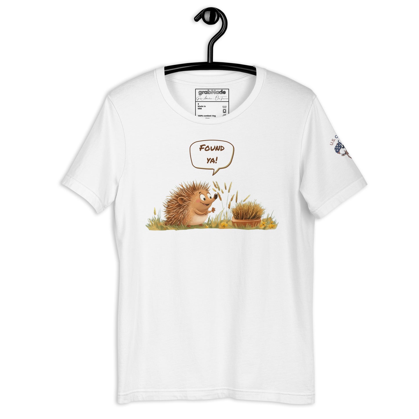 Product mockup. White T-shirt on hanger featuring a smiling cartoon hedgehog in grass saying "Found ya!" with playful, earthy colors.