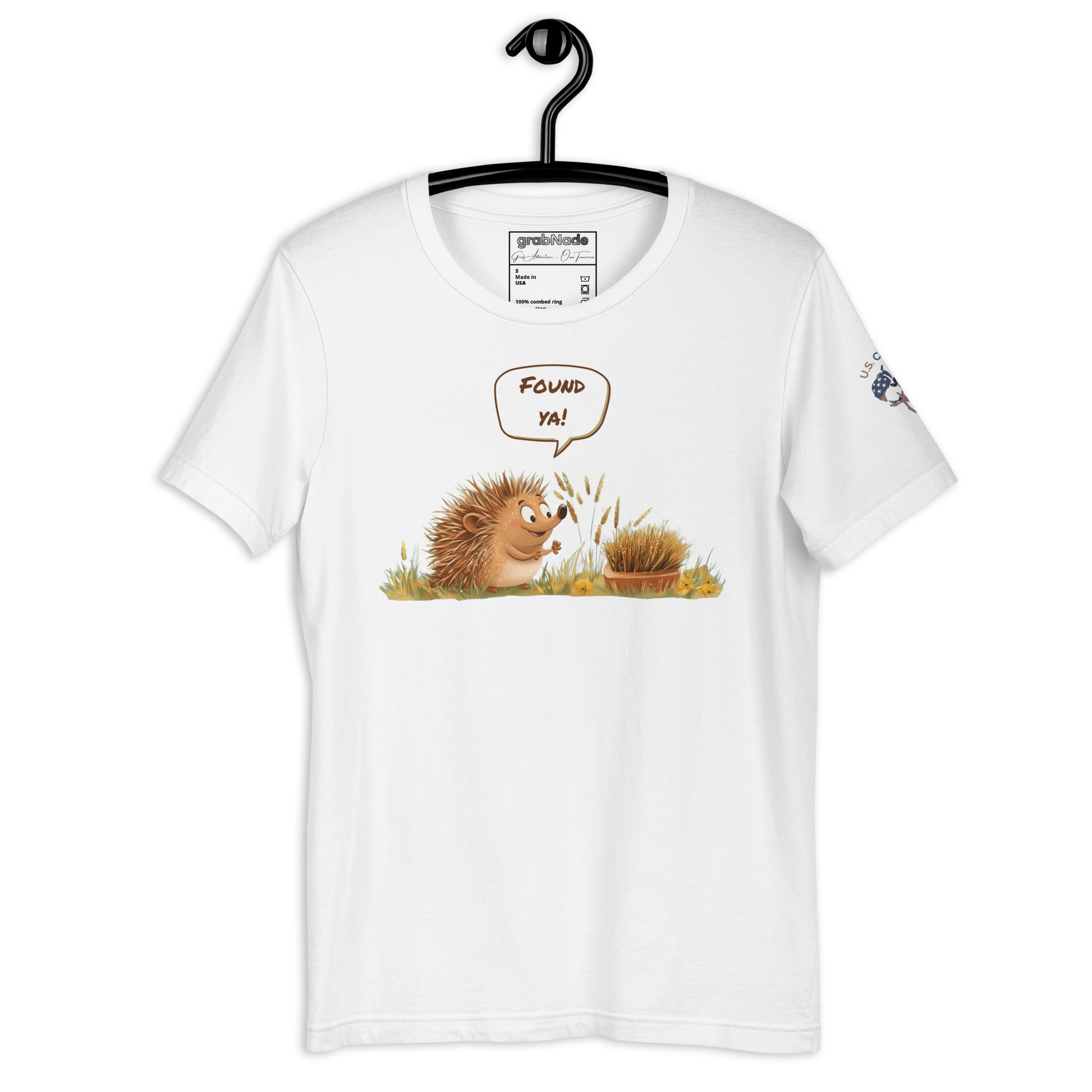 Product mockup. White T-shirt on hanger featuring a smiling cartoon hedgehog in grass saying "Found ya!" with playful, earthy colors.
