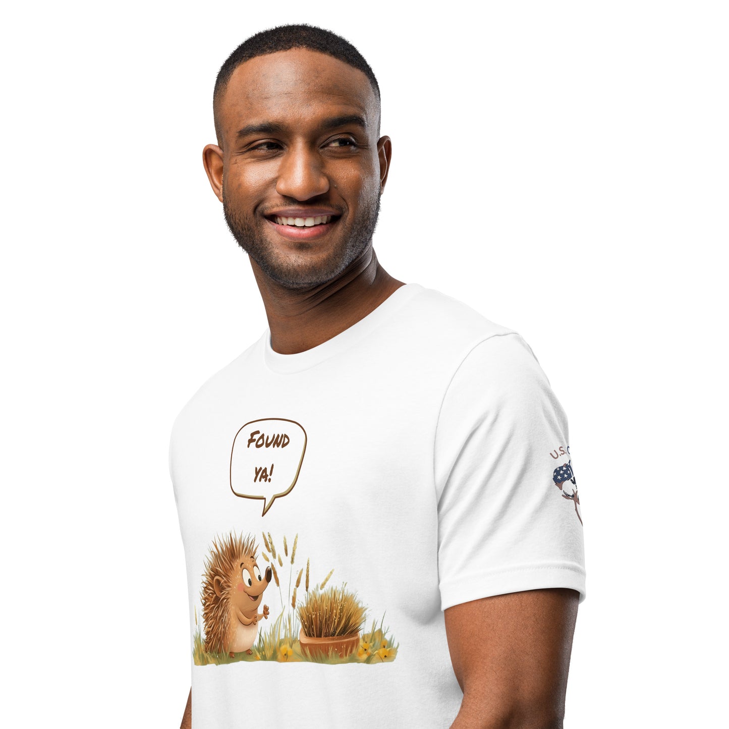 Product mockup. Man in white T-shirt featuring a smiling cartoon hedgehog exclaiming "Found ya!" beside a bush and yellow flowers.