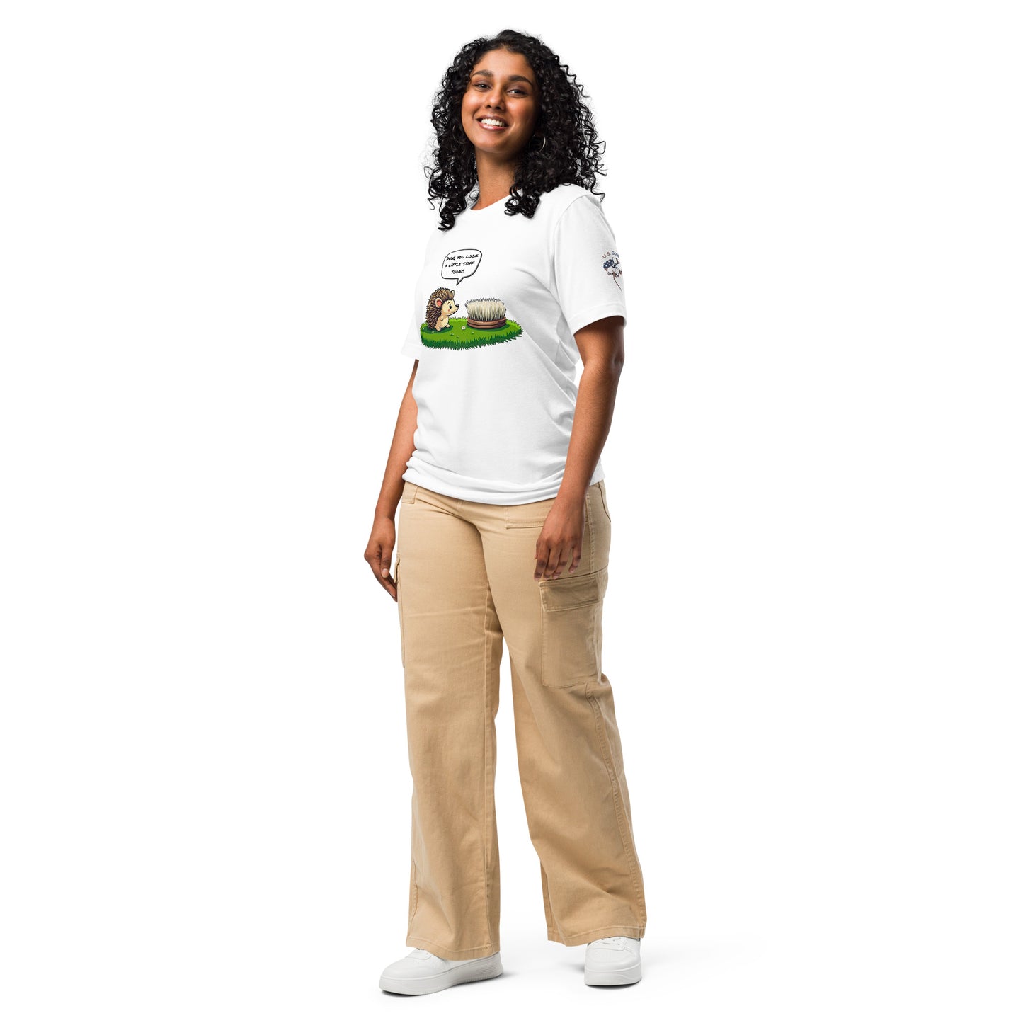 Product mockup. A woman dressed in beige pants and a white T-shirt with a cartoon hedgehog remarking “Dude, you look a little stiff today!” to a brush, standing in a full-body pose.