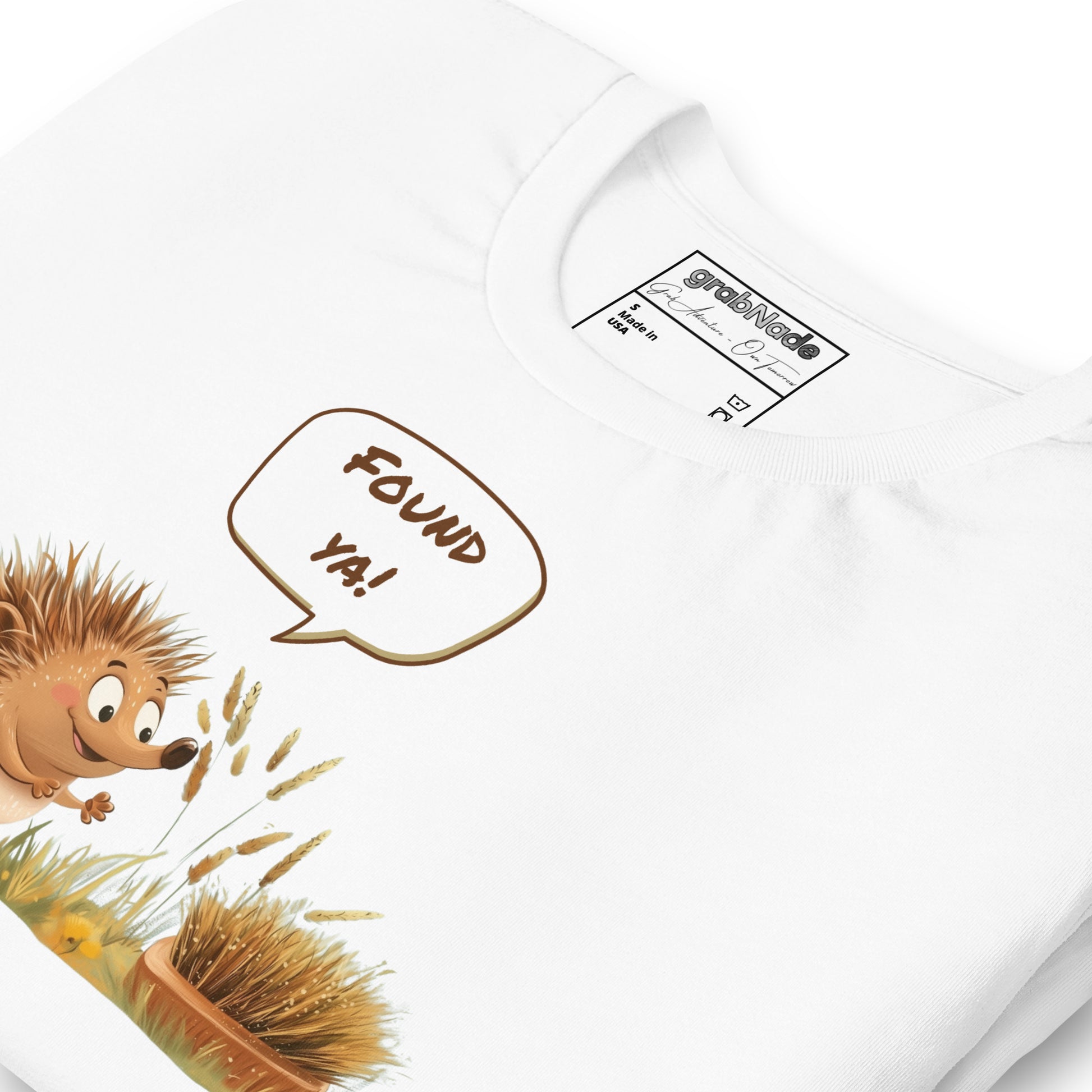 Product mockup. Close-up of a white T-shirt featuring a smiling cartoon hedgehog exclaiming “Found ya!” toward a brush disguised as a hedgehog in grass.