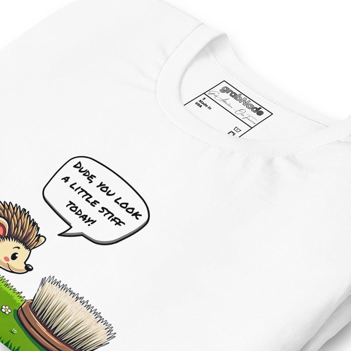 Product mockup. Close-up of a white T-shirt with a cartoon hedgehog saying, “DUDE, YOU LOOK A LITTLE STIFF TODAY!” to a round brush in grass.