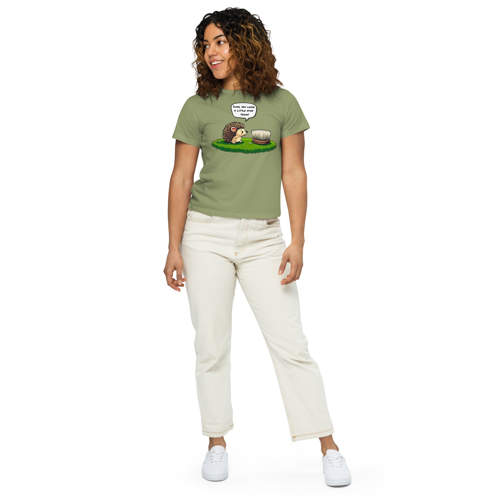 Product mockup. Full-body view of a woman in a light green high-waisted t-shirt with a whimsical hedgehog and scrubbing brush cartoon print. She wears off-white pants and white sneakers, standing against a plain backdrop and looking off to the side.