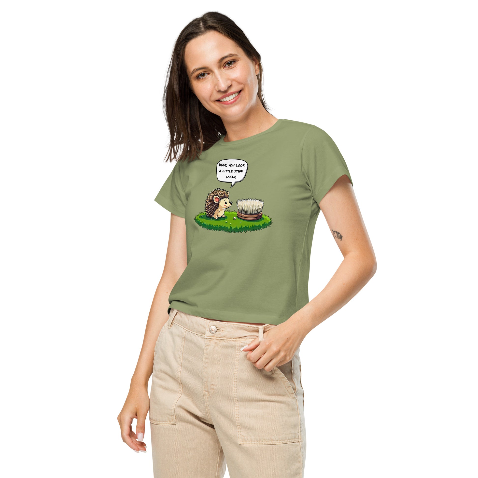 Product mockup. Front view of a woman wearing a light green high-waisted t-shirt with a cartoon design. The graphic shows a cheerful hedgehog facing a scrub brush on grass, with a speech bubble that says, 'Dude, you look a little stiff today!' She is smiling and has one hand in her beige pants pocket, standing against a plain background.