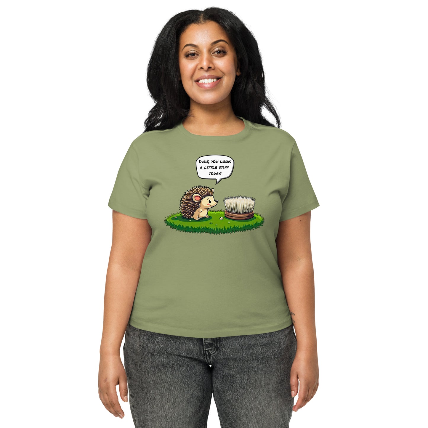 Product mockup. Front view of a woman wearing a light green high-waisted t-shirt with a playful cartoon print. The graphic features a smiling hedgehog facing a scrubbing brush on a patch of green grass, with the speech bubble, 'Dude, you look a little stiff today!' She stands against a plain background