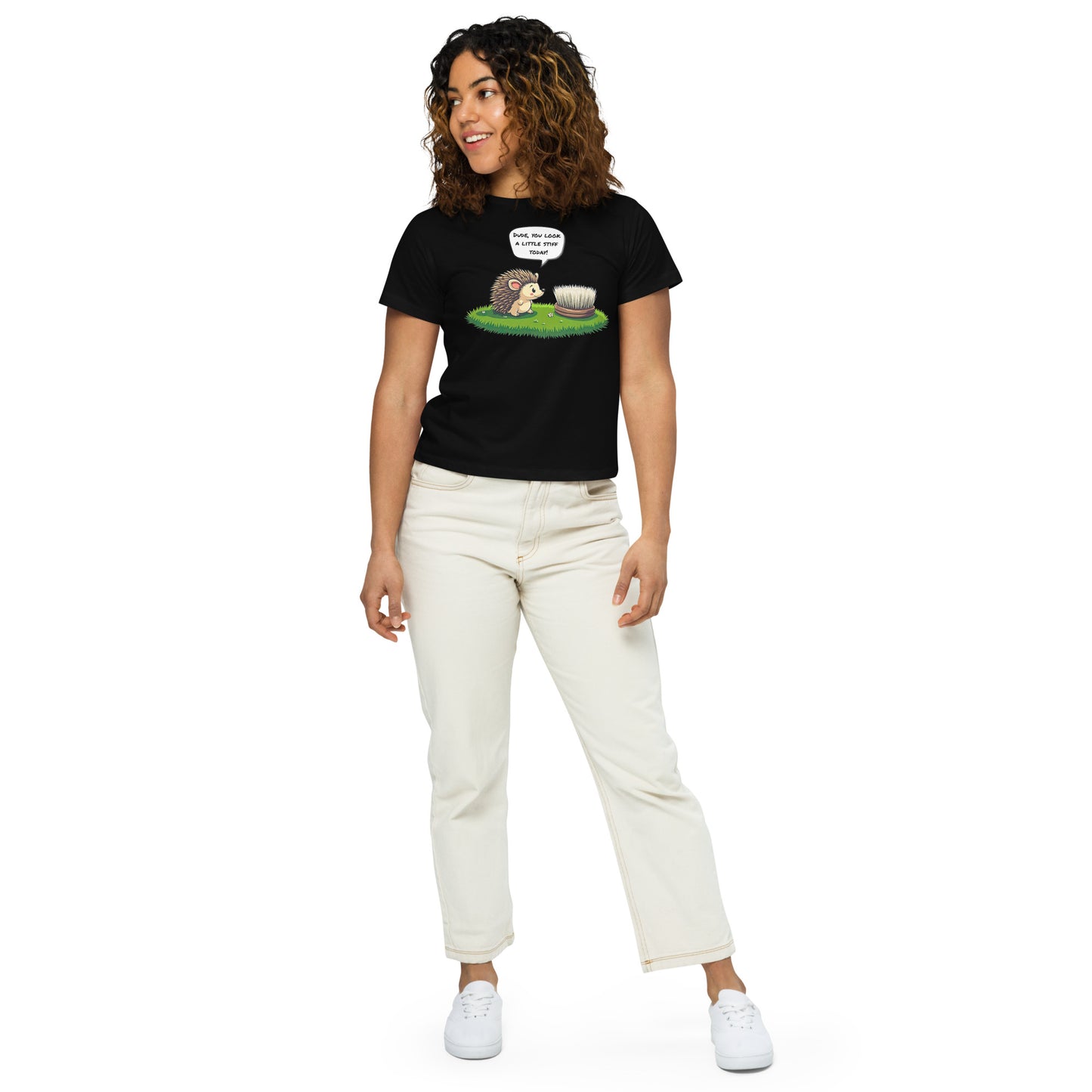 Product mockup. Full-body view of a woman in a black high-waisted t-shirt with a whimsical hedgehog and scrubbing brush cartoon print. She wears off-white pants and white sneakers, standing against a plain backdrop and looking off to the side.