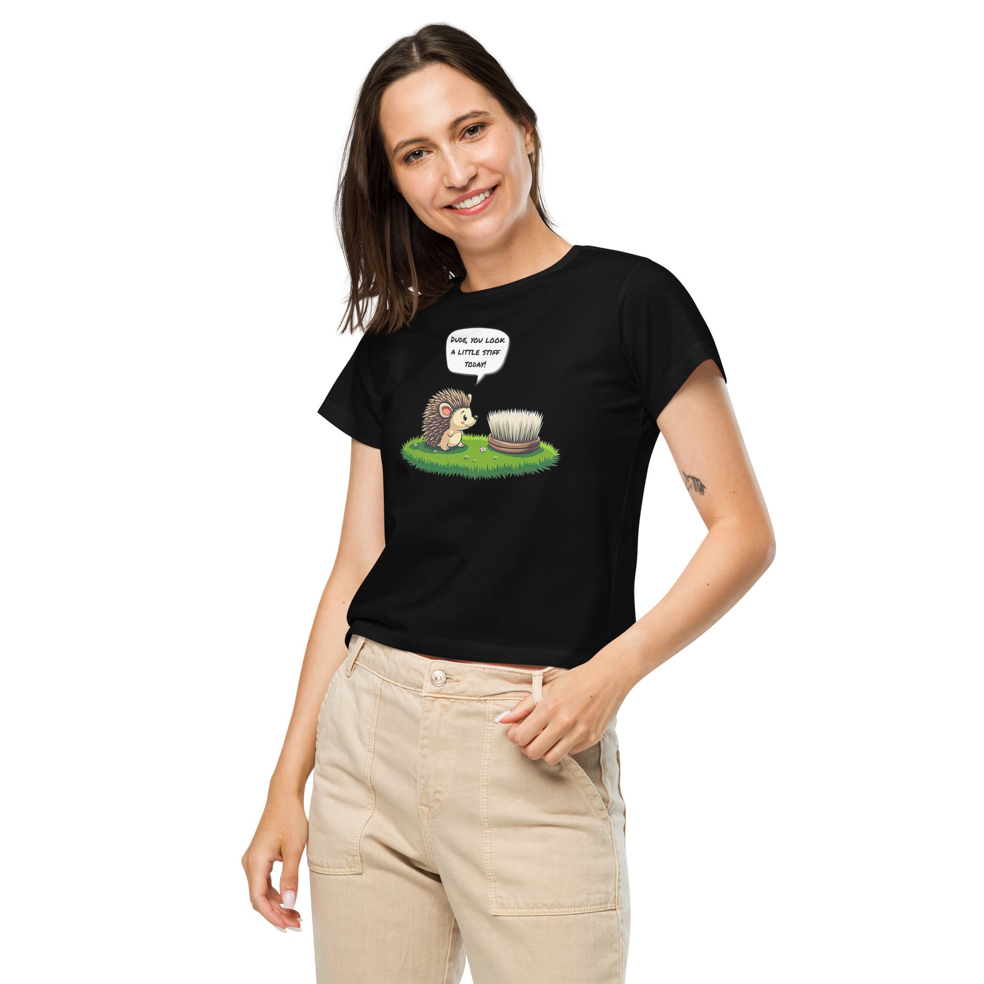 Product mockup. Front view of a woman wearing a black high-waisted t-shirt with a cartoon design. The graphic shows a cheerful hedgehog facing a scrub brush on grass, with a speech bubble that says, 'Dude, you look a little stiff today!' She is smiling and has one hand in her beige pants pocket, standing against a plain background.