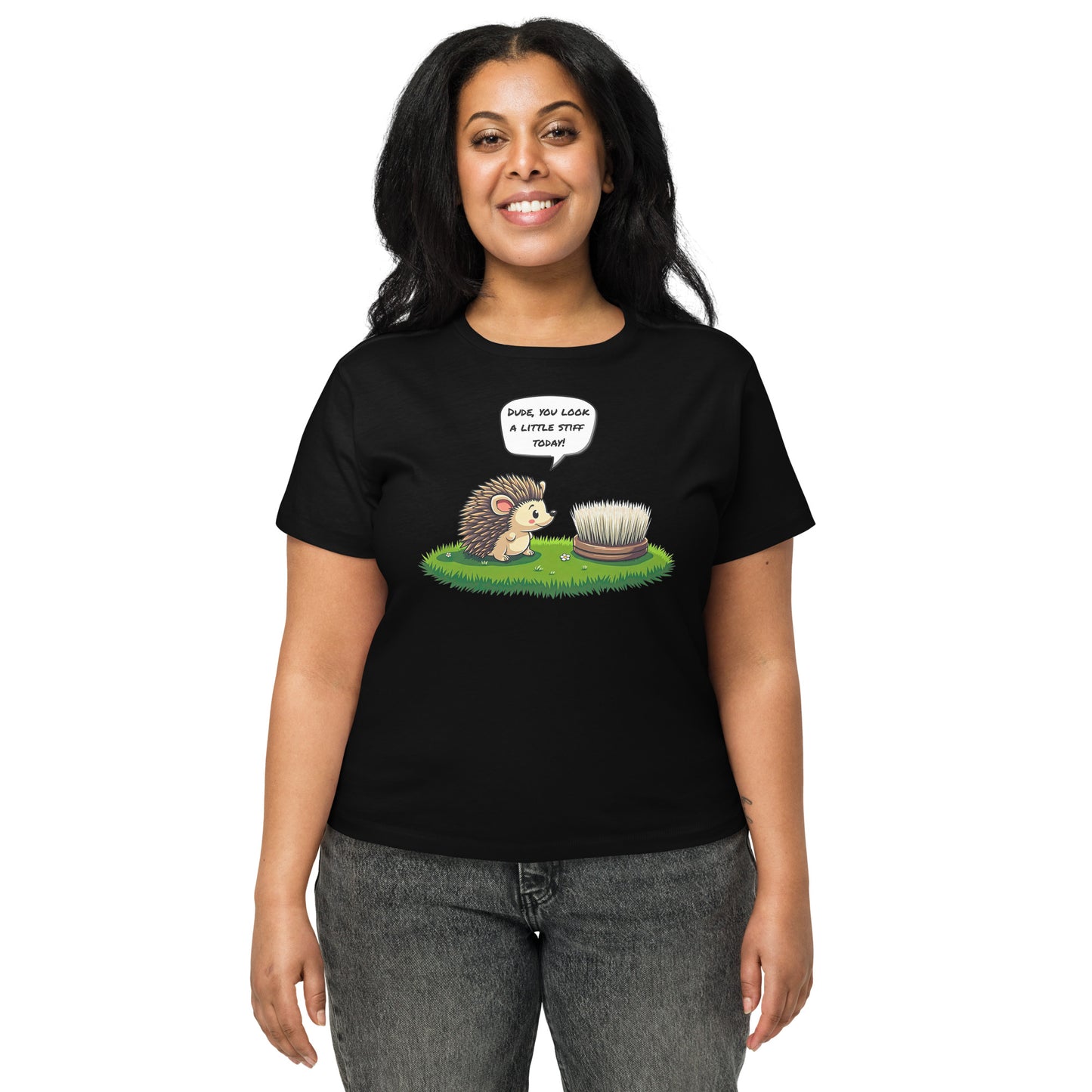 Product mockup. Front view of a woman wearing a black high-waisted t-shirt with a playful cartoon print. The graphic features a smiling hedgehog facing a scrubbing brush on a patch of green grass, with the speech bubble, 'Dude, you look a little stiff today!' She stands against a plain background