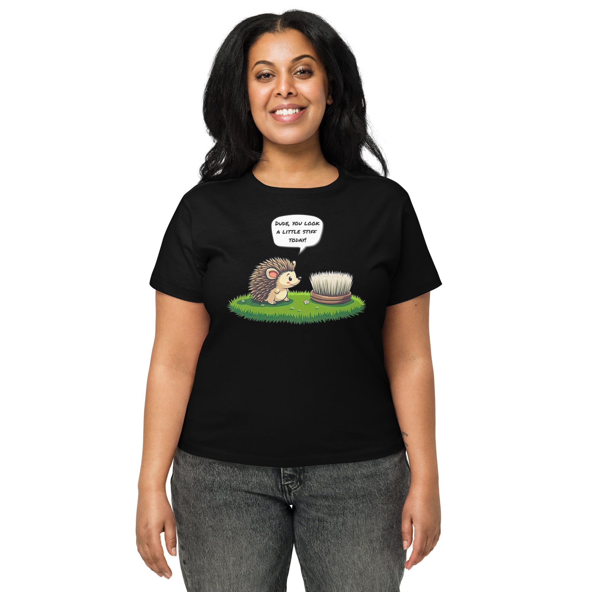 Product mockup. Front view of a woman wearing a black high-waisted t-shirt with a playful cartoon print. The graphic features a smiling hedgehog facing a scrubbing brush on a patch of green grass, with the speech bubble, 'Dude, you look a little stiff today!' She stands against a plain background
