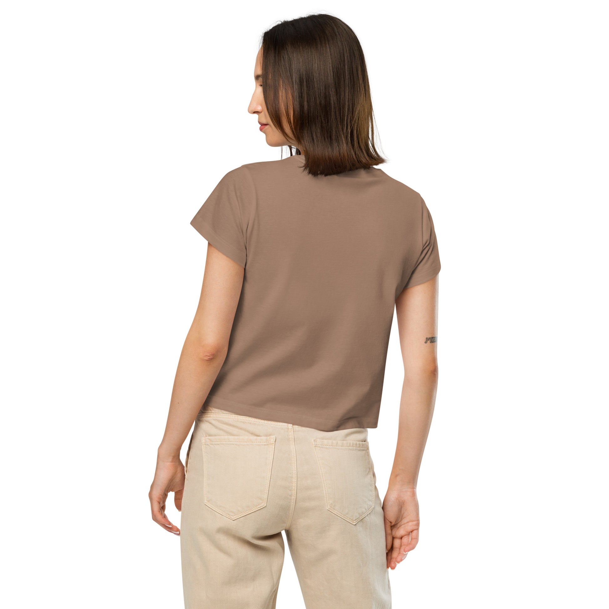Product mockup. Back view of a woman wearing a latte high-waisted t-shirt and beige pants. The back of the t-shirt is plain, and she is facing away from the camera, standing against a simple white background.