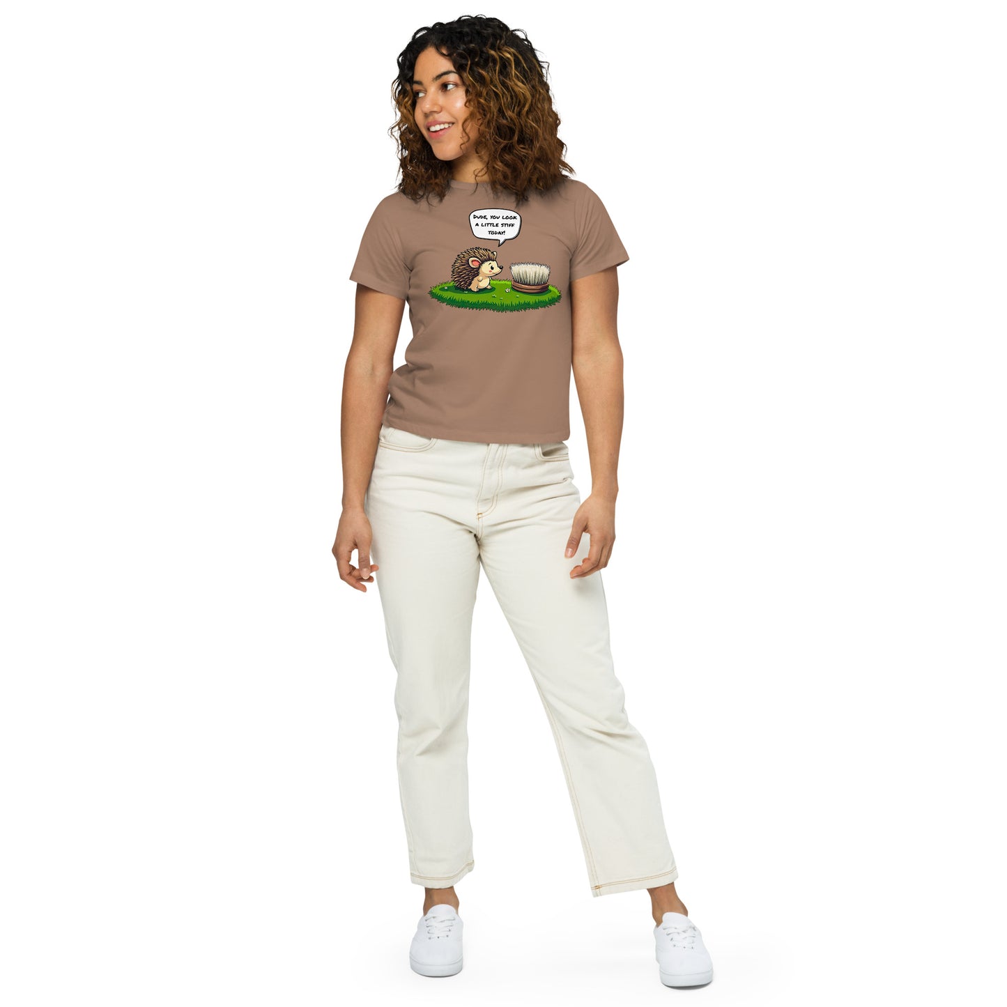 Product mockup. Full-body view of a woman in a latte high-waisted t-shirt with a whimsical hedgehog and scrubbing brush cartoon print. She wears off-white pants and white sneakers, standing against a plain backdrop and looking off to the side.