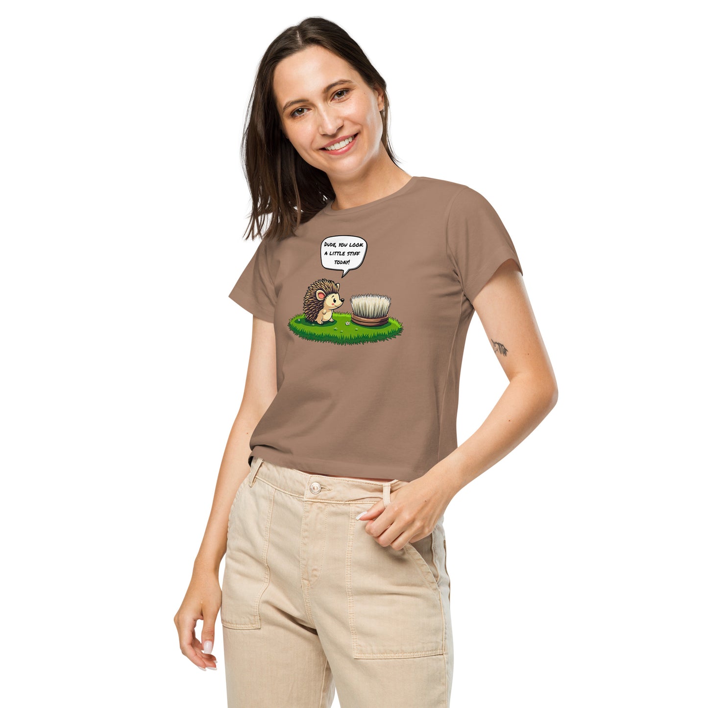 Product mockup. Front view of a woman wearing a latte high-waisted t-shirt with a cartoon design. The graphic shows a cheerful hedgehog facing a scrub brush on grass, with a speech bubble that says, 'Dude, you look a little stiff today!' She is smiling and has one hand in her beige pants pocket, standing against a plain background.