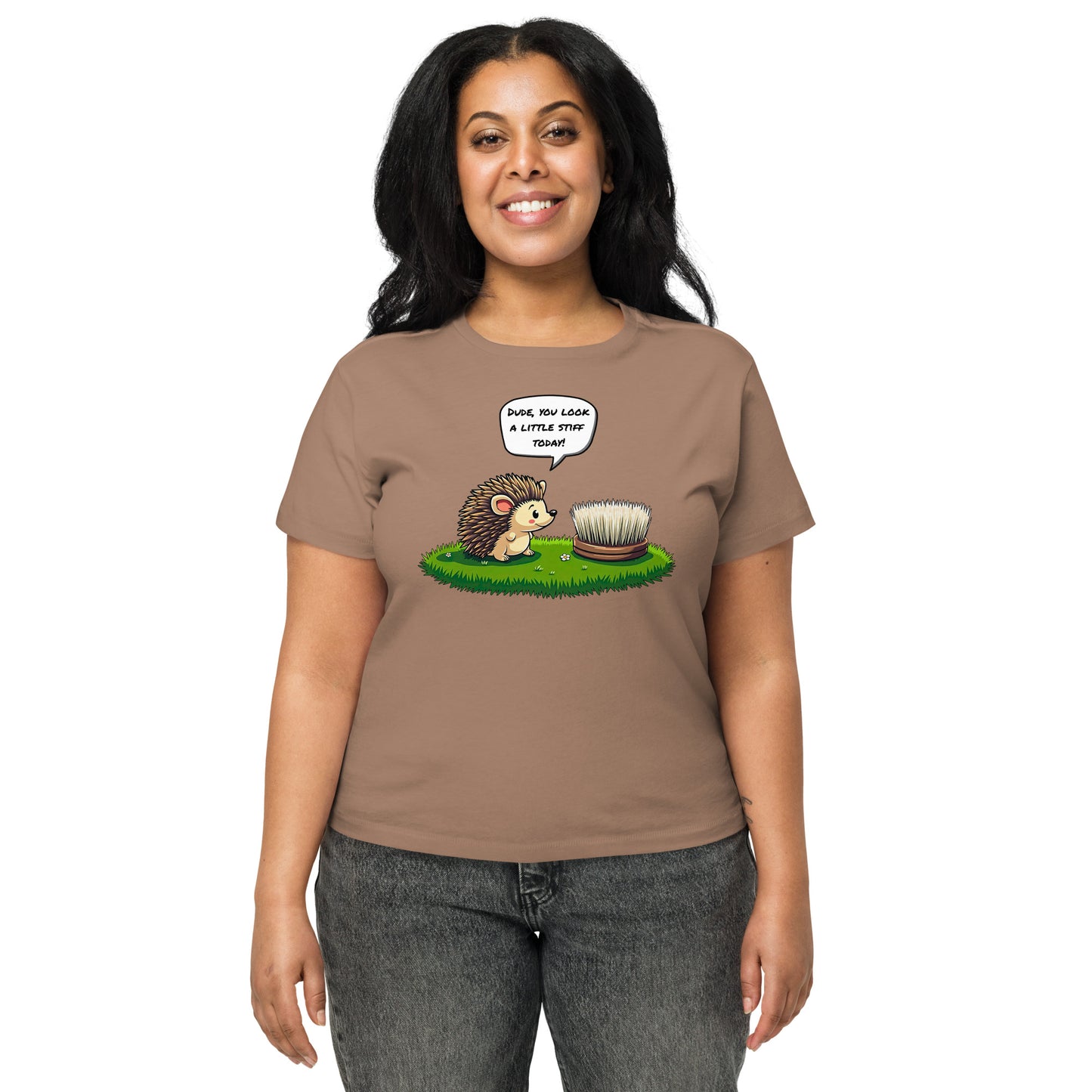 Product mockup. Front view of a woman wearing a latte high-waisted t-shirt with a playful cartoon print. The graphic features a smiling hedgehog facing a scrubbing brush on a patch of green grass, with the speech bubble, 'Dude, you look a little stiff today!' She stands against a plain background