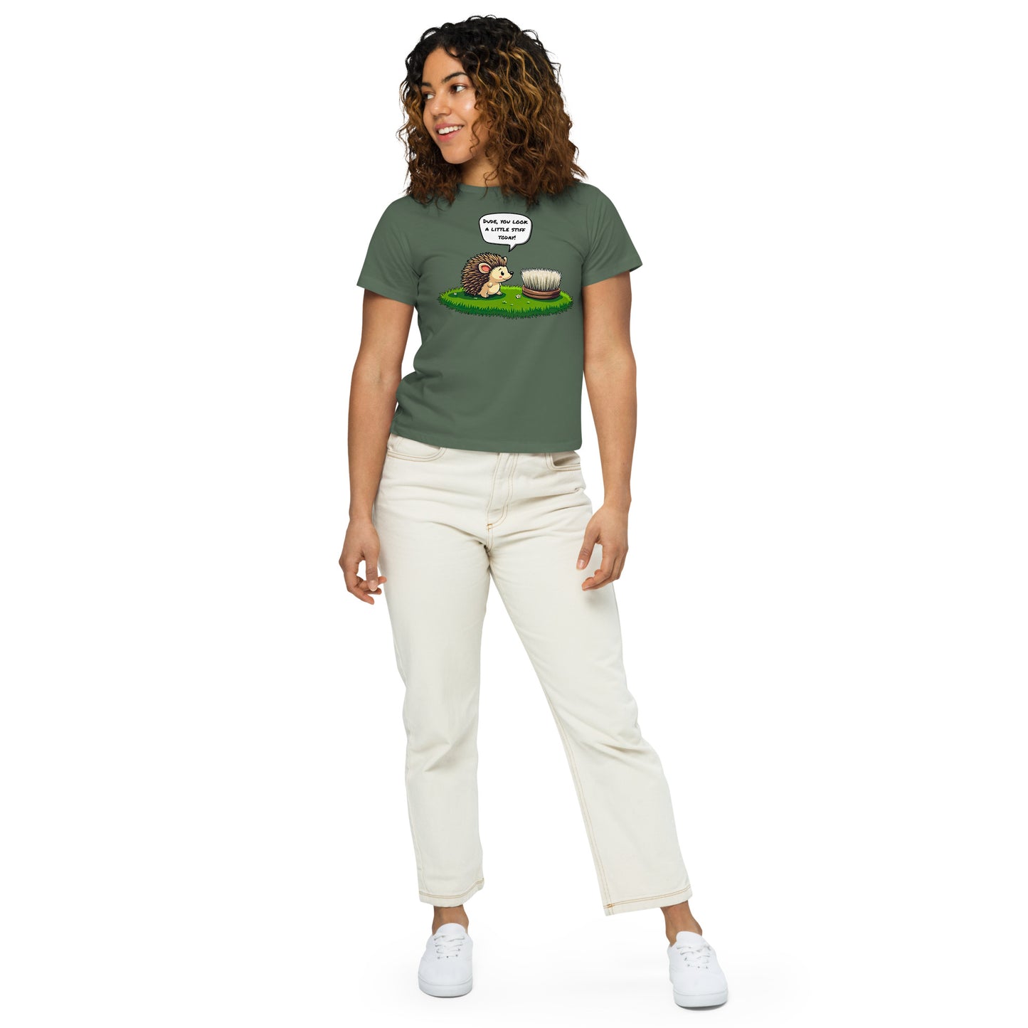 Product mockup. Full-body view of a woman in a green pine high-waisted t-shirt with a whimsical hedgehog and scrubbing brush cartoon print. She wears off-white pants and white sneakers, standing against a plain backdrop and looking off to the side.