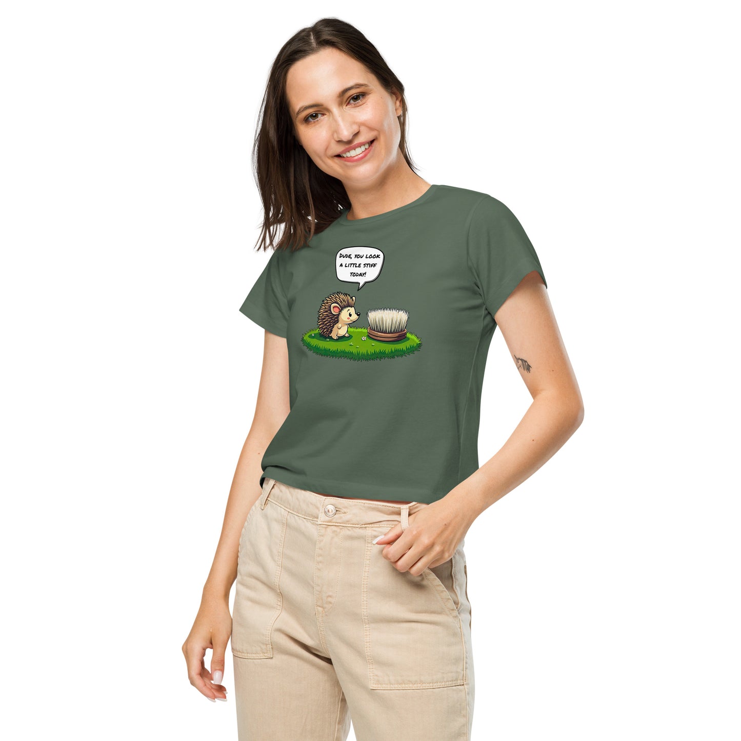 Product mockup. Front view of a woman wearing a green pine high-waisted t-shirt with a cartoon design. The graphic shows a cheerful hedgehog facing a scrub brush on grass, with a speech bubble that says, 'Dude, you look a little stiff today!' She is smiling and has one hand in her beige pants pocket, standing against a plain background.