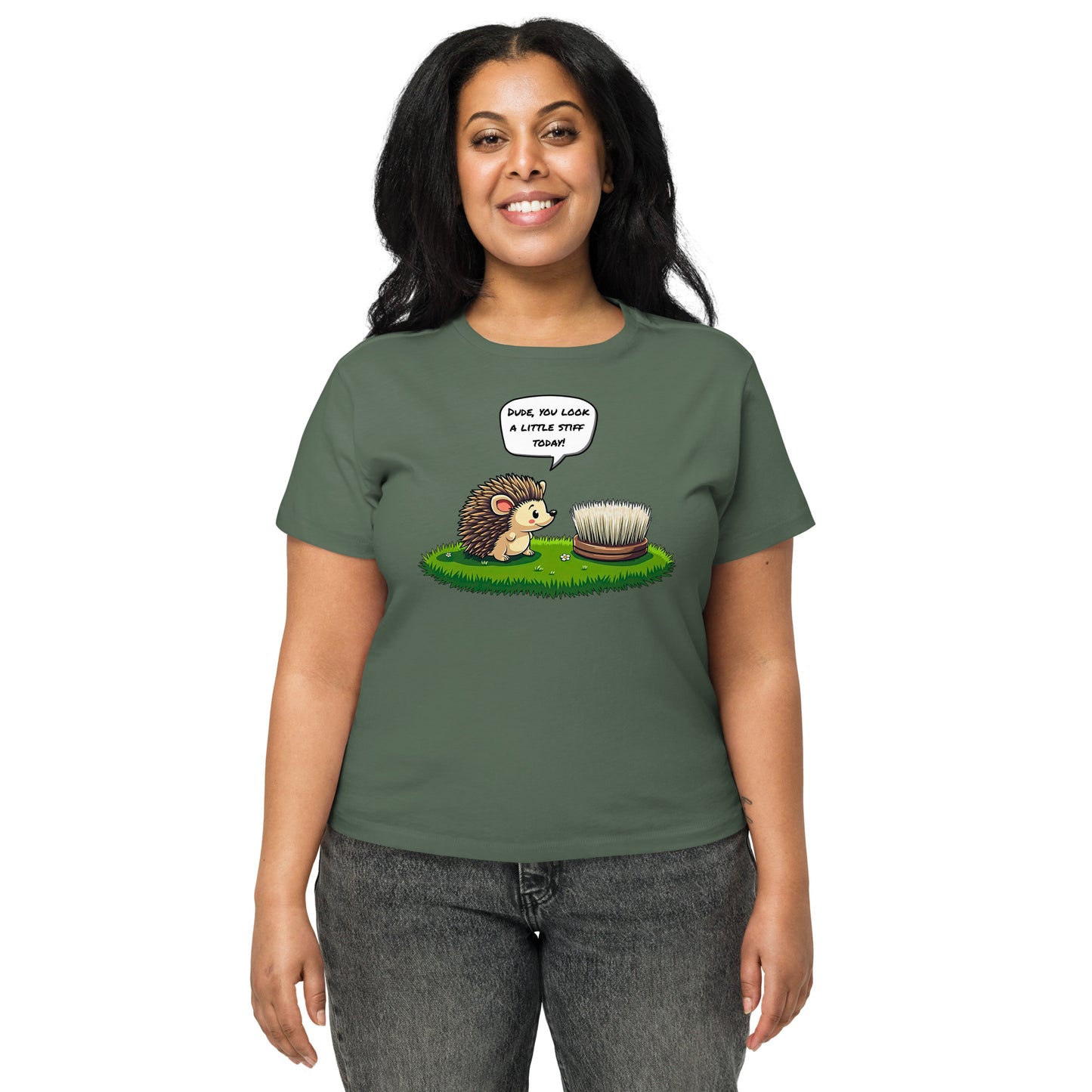 Product mockup. Front view of a woman wearing a green pine high-waisted t-shirt with a playful cartoon print. The graphic features a smiling hedgehog facing a scrubbing brush on a patch of green grass, with the speech bubble, 'Dude, you look a little stiff today!' She stands against a plain background