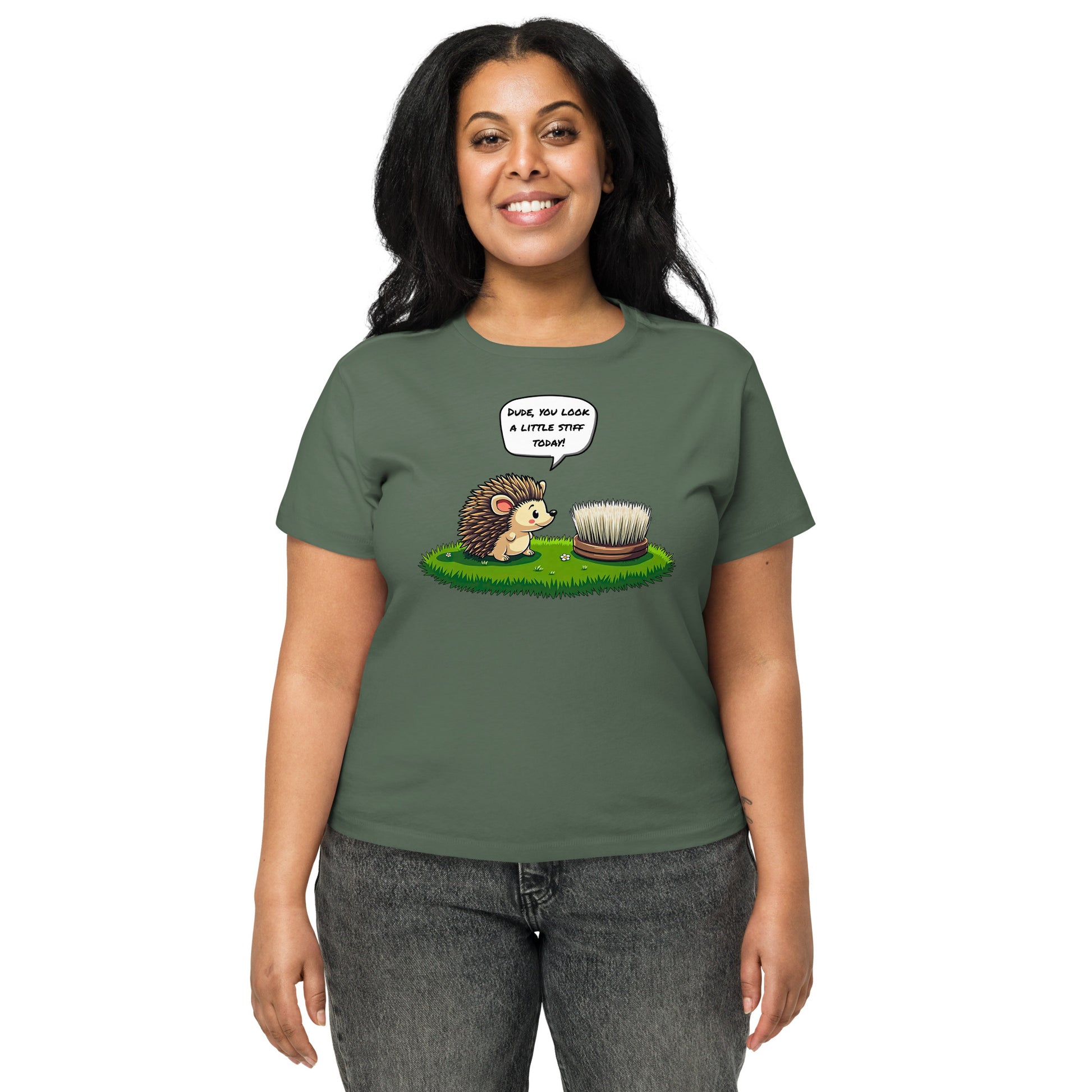 Product mockup. Front view of a woman wearing a green pine high-waisted t-shirt with a playful cartoon print. The graphic features a smiling hedgehog facing a scrubbing brush on a patch of green grass, with the speech bubble, 'Dude, you look a little stiff today!' She stands against a plain background