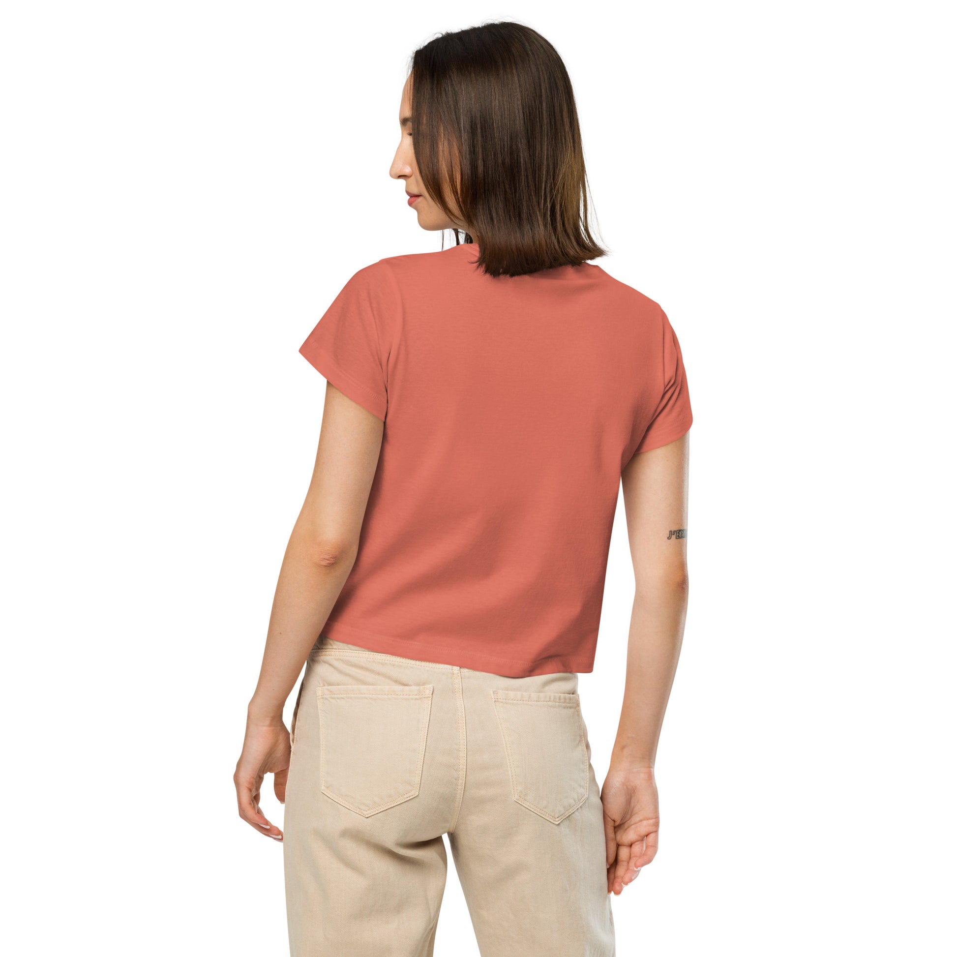 Product mockup. Back view of a woman wearing a red sorbet high-waisted t-shirt and beige pants. The back of the t-shirt is plain, and she is facing away from the camera, standing against a simple white background.