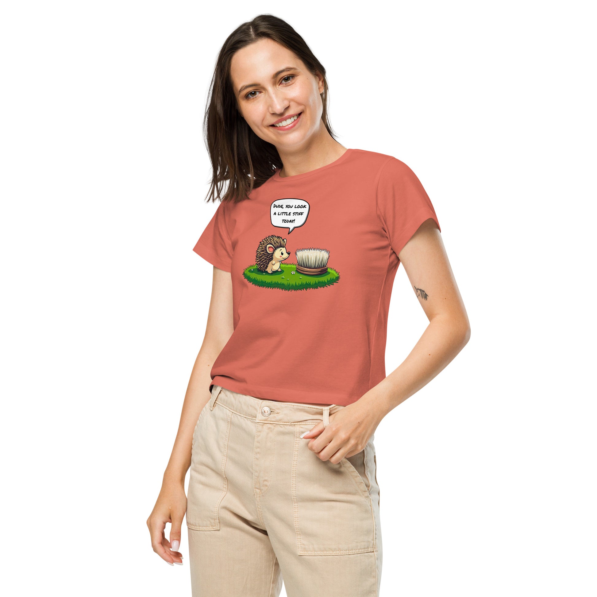 Product mockup. Front view of a woman wearing a red sorbet high-waisted t-shirt with a cartoon design. The graphic shows a cheerful hedgehog facing a scrub brush on grass, with a speech bubble that says, 'Dude, you look a little stiff today!' She is smiling and has one hand in her beige pants pocket, standing against a plain background.