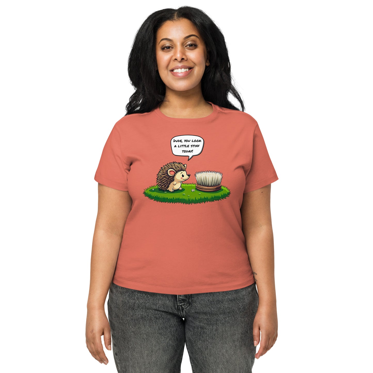 Product mockup. Front view of a woman wearing a red sorbet high-waisted t-shirt with a playful cartoon print. The graphic features a smiling hedgehog facing a scrubbing brush on a patch of green grass, with the speech bubble, 'Dude, you look a little stiff today!' She stands against a plain background
