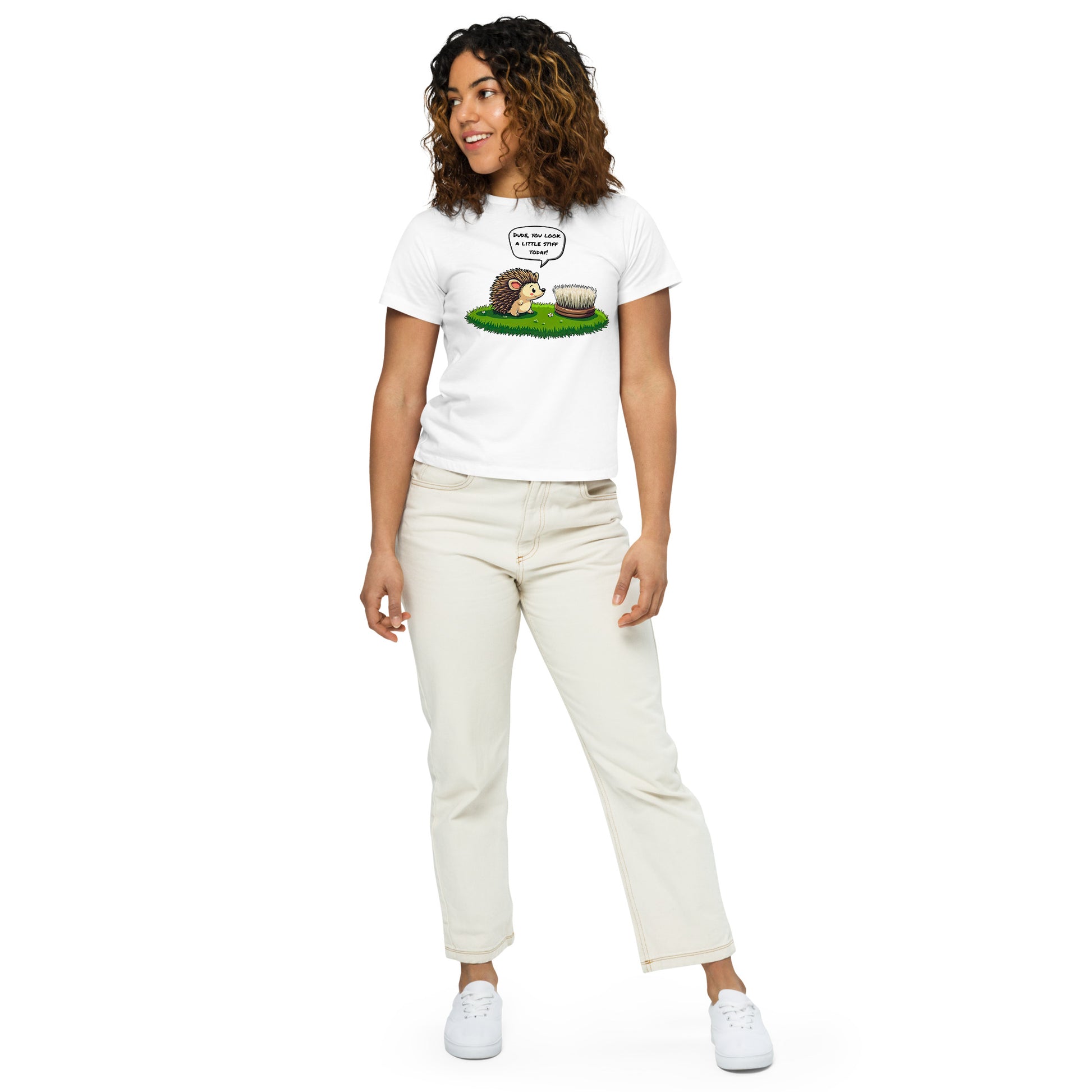 Product mockup. Full-body view of a woman in a white high-waisted t-shirt with a whimsical hedgehog and scrubbing brush cartoon print. She wears off-white pants and white sneakers, standing against a plain backdrop and looking off to the side.