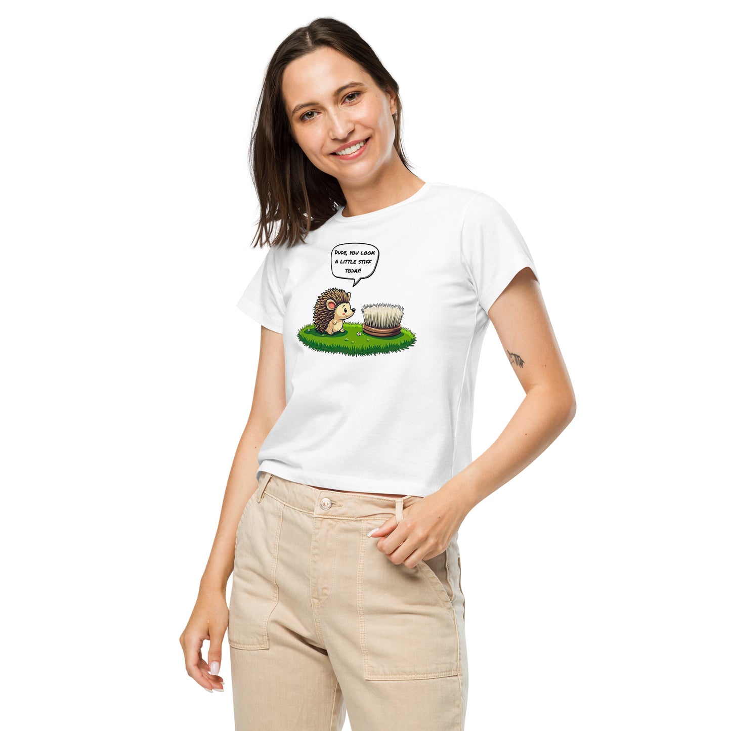Product mockup. Front view of a woman wearing a white high-waisted t-shirt with a cartoon design. The graphic shows a cheerful hedgehog facing a scrub brush on grass, with a speech bubble that says, 'Dude, you look a little stiff today!' She is smiling and has one hand in her beige pants pocket, standing against a plain background.