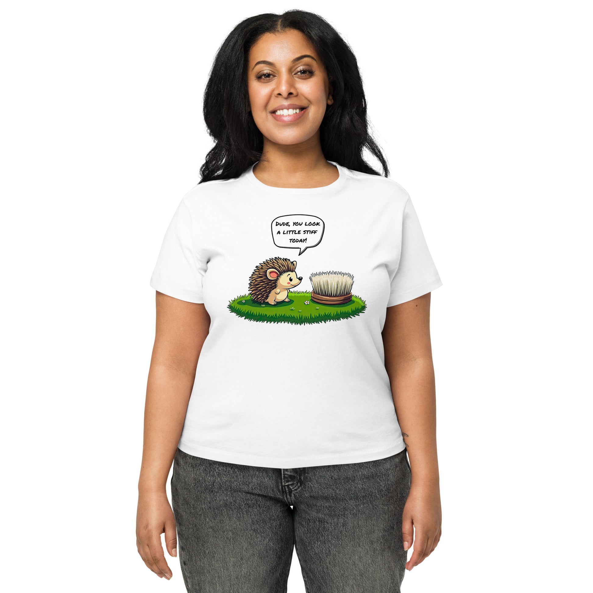 Product mockup. Front view of a woman wearing a white high-waisted t-shirt with a playful cartoon print. The graphic features a smiling hedgehog facing a scrubbing brush on a patch of green grass, with the speech bubble, 'Dude, you look a little stiff today!' She stands against a plain background