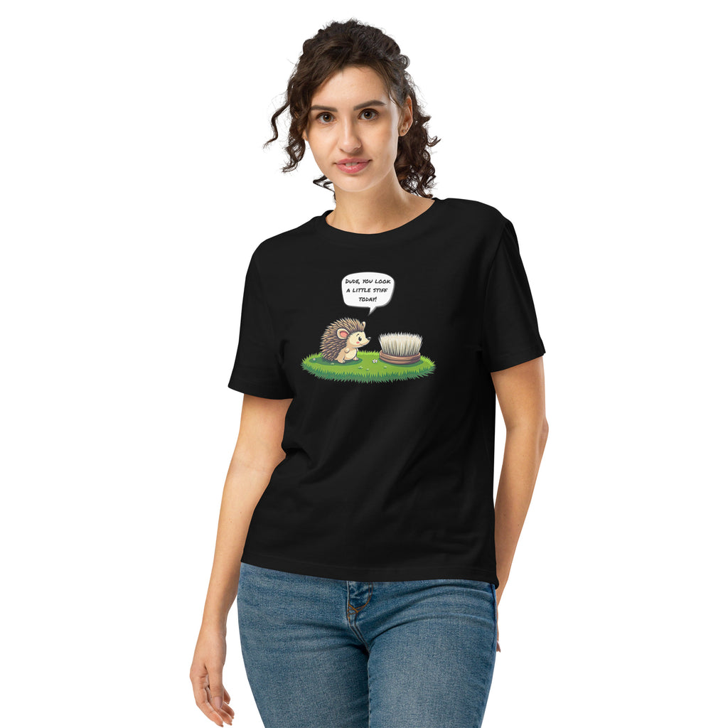 Product mockup. A happy woman wearing a black organic cotton t-shirt featuring a playful cartoon illustration of a hedgehog standing on green grass beside a scrubbing brush. The hedgehog has a speech bubble that says, 'DUDE, YOU LOOK A LITTLE STIFF TODAY!'
