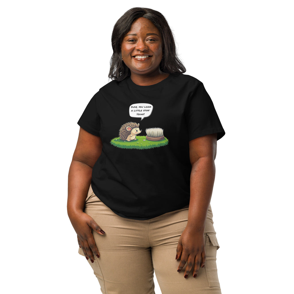 Product mockup. A happy woman wearing a 2XL black organic cotton t-shirt featuring a playful cartoon illustration of a hedgehog standing on green grass beside a scrubbing brush. The hedgehog has a speech bubble that says, 'DUDE, YOU LOOK A LITTLE STIFF TODAY!'
