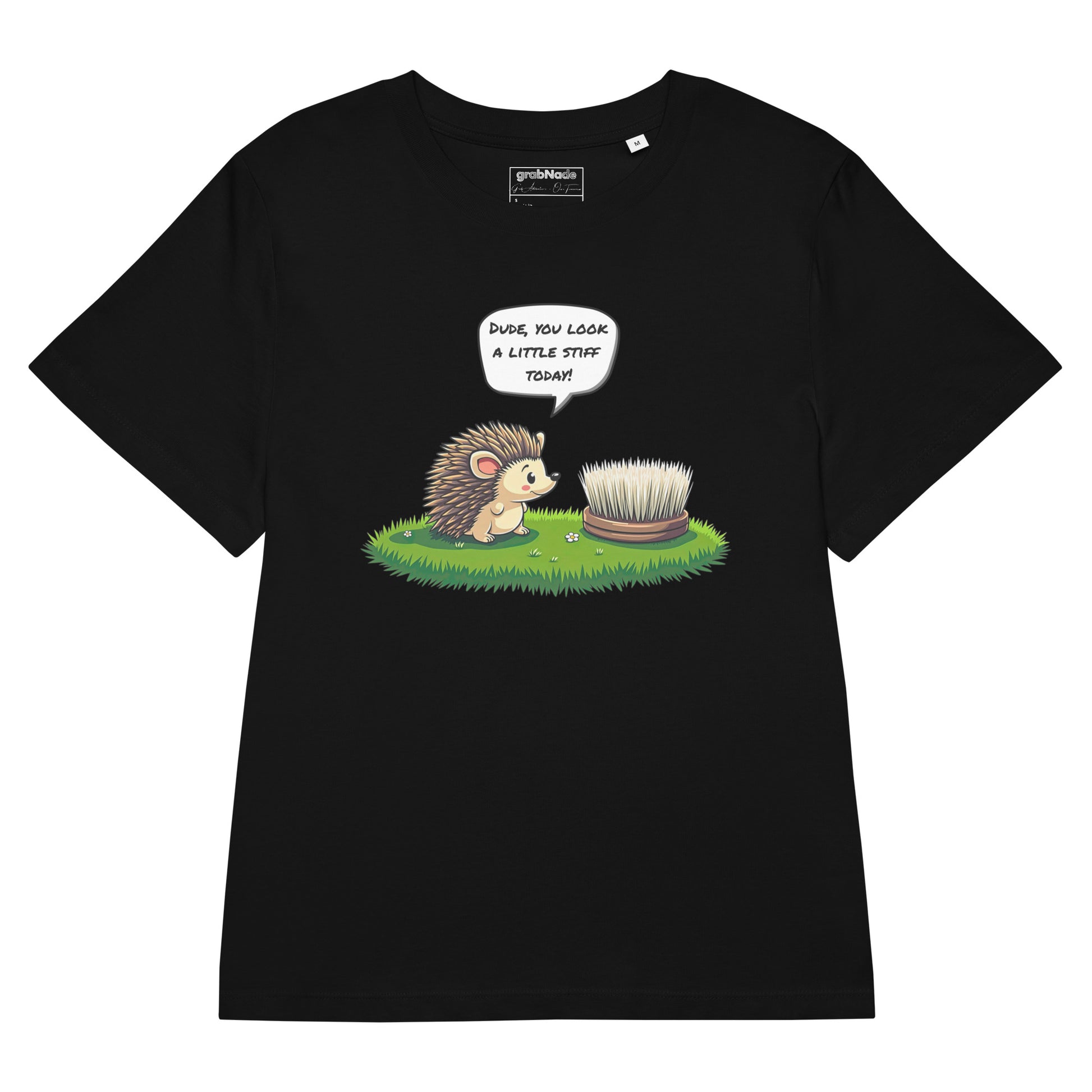 Product mockup. A flat layout black t-shirt made from organic cotton, featuring a playful illustration on the front. The design shows a cute cartoon hedgehog standing on green grass, looking at a stiff scrubbing brush, with a speech bubble that reads, 'DUDE, YOU LOOK A LITTLE STIFF TODAY!' The 'grabNade' brand label is visible inside the neckline.