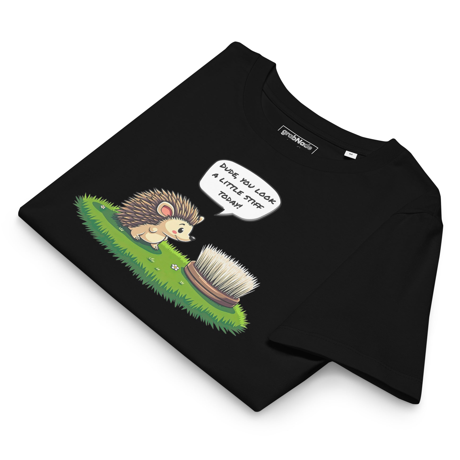 Product mockup. A folded black t-shirt made from organic cotton, featuring a playful illustration on the front. The design shows a cute cartoon hedgehog standing on green grass, looking at a stiff scrubbing brush, with a speech bubble that reads, 'DUDE, YOU LOOK A LITTLE STIFF TODAY!' The 'grabNade' brand label is visible inside the neckline.