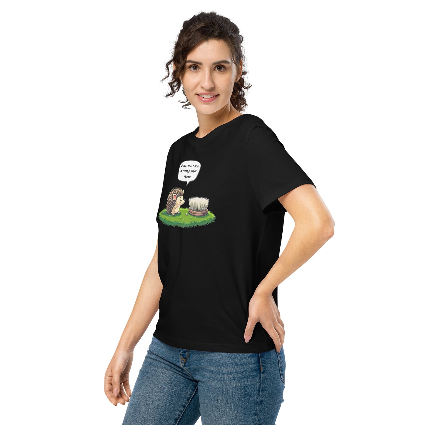 Product mockup. A happy woman wearing a black organic cotton t-shirt featuring a playful cartoon illustration of a hedgehog standing on green grass beside a scrubbing brush. The hedgehog has a speech bubble that says, 'DUDE, YOU LOOK A LITTLE STIFF TODAY!'