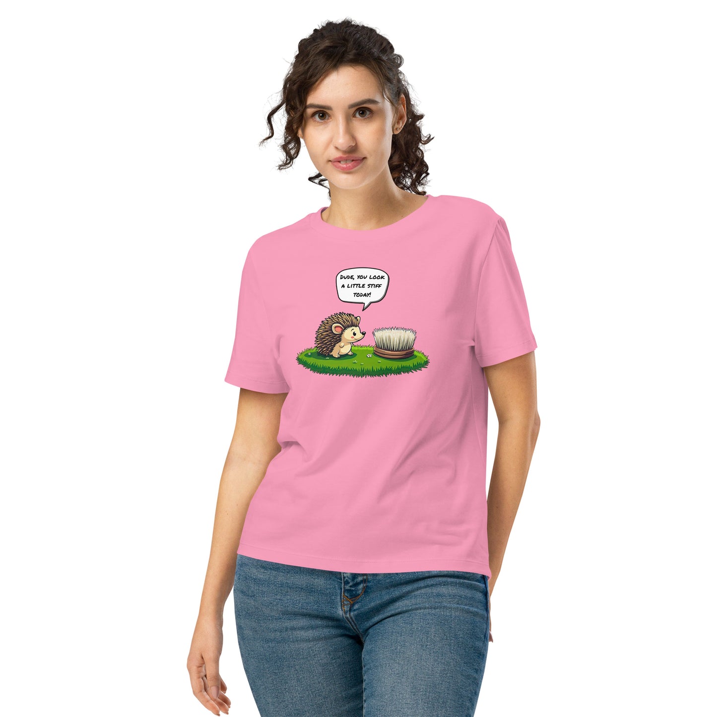 Product mockup. A happy woman wearing a pink organic cotton t-shirt featuring a playful cartoon illustration of a hedgehog standing on green grass beside a scrubbing brush. The hedgehog has a speech bubble that says, 'DUDE, YOU LOOK A LITTLE STIFF TODAY!'