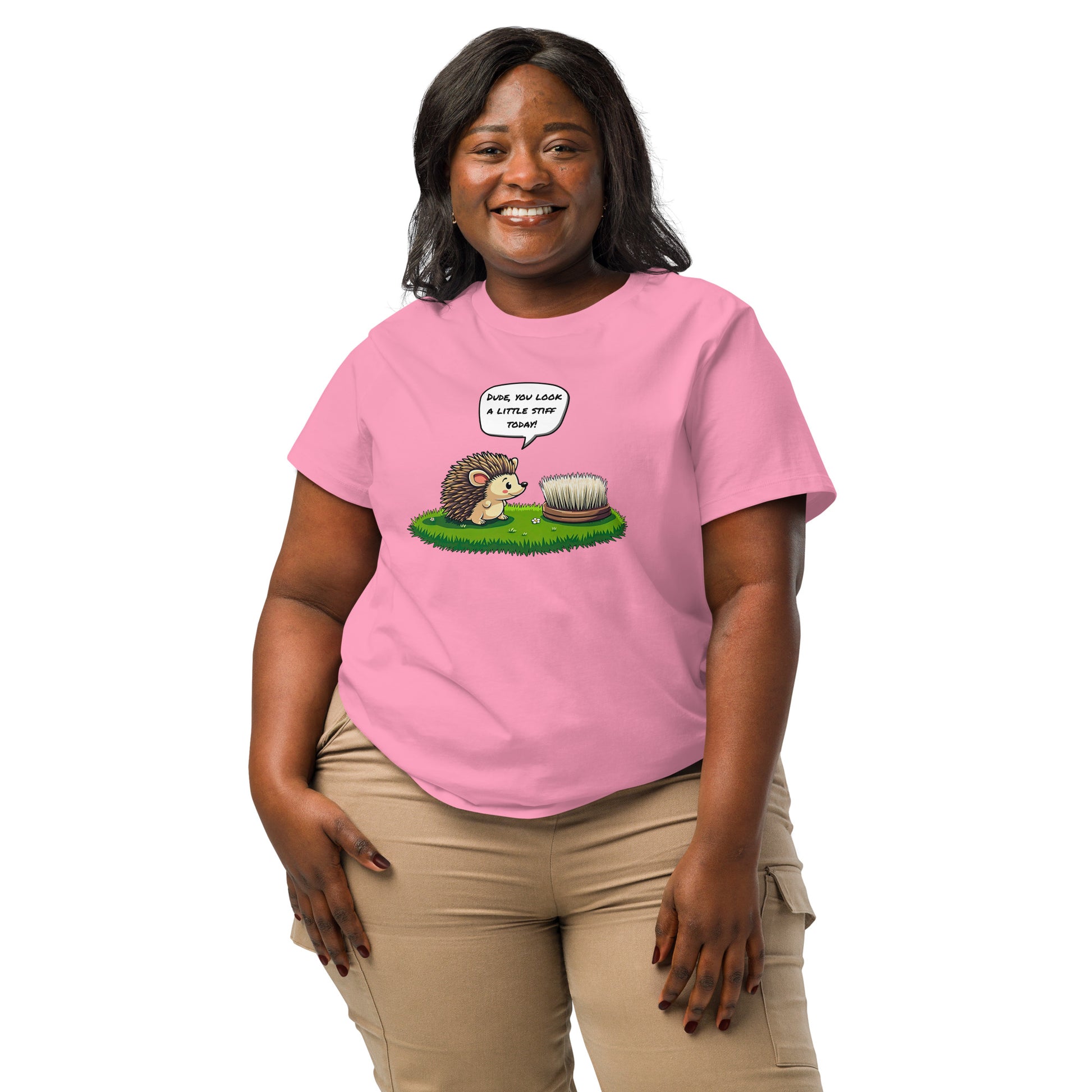 Product mockup. A happy woman wearing a 2XL pink organic cotton t-shirt featuring a playful cartoon illustration of a hedgehog standing on green grass beside a scrubbing brush. The hedgehog has a speech bubble that says, 'DUDE, YOU LOOK A LITTLE STIFF TODAY!'