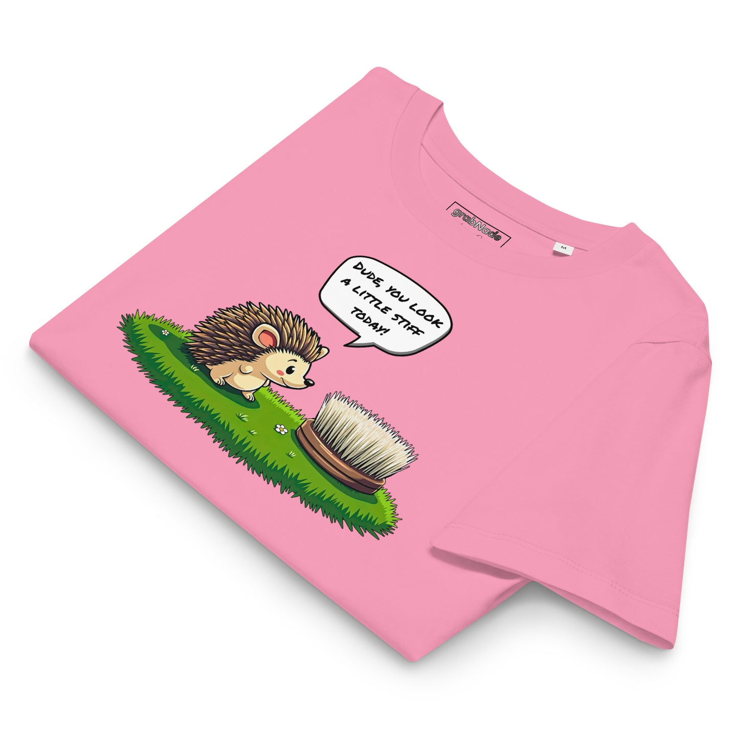Product mockup. A folded pink t-shirt made from organic cotton, featuring a playful illustration on the front. The design shows a cute cartoon hedgehog standing on green grass, looking at a stiff scrubbing brush, with a speech bubble that reads, 'DUDE, YOU LOOK A LITTLE STIFF TODAY!' The 'grabNade' brand label is visible inside the neckline.