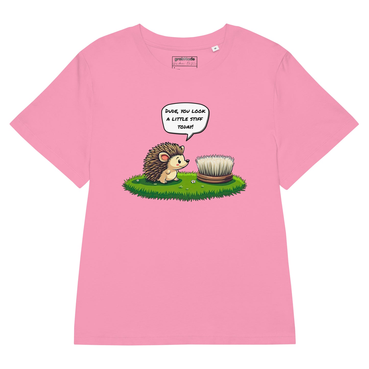 Product mockup. A flat layout pink t-shirt made from organic cotton, featuring a playful illustration on the front. The design shows a cute cartoon hedgehog standing on green grass, looking at a stiff scrubbing brush, with a speech bubble that reads, 'DUDE, YOU LOOK A LITTLE STIFF TODAY!' The 'grabNade' brand label is visible inside the neckline.
