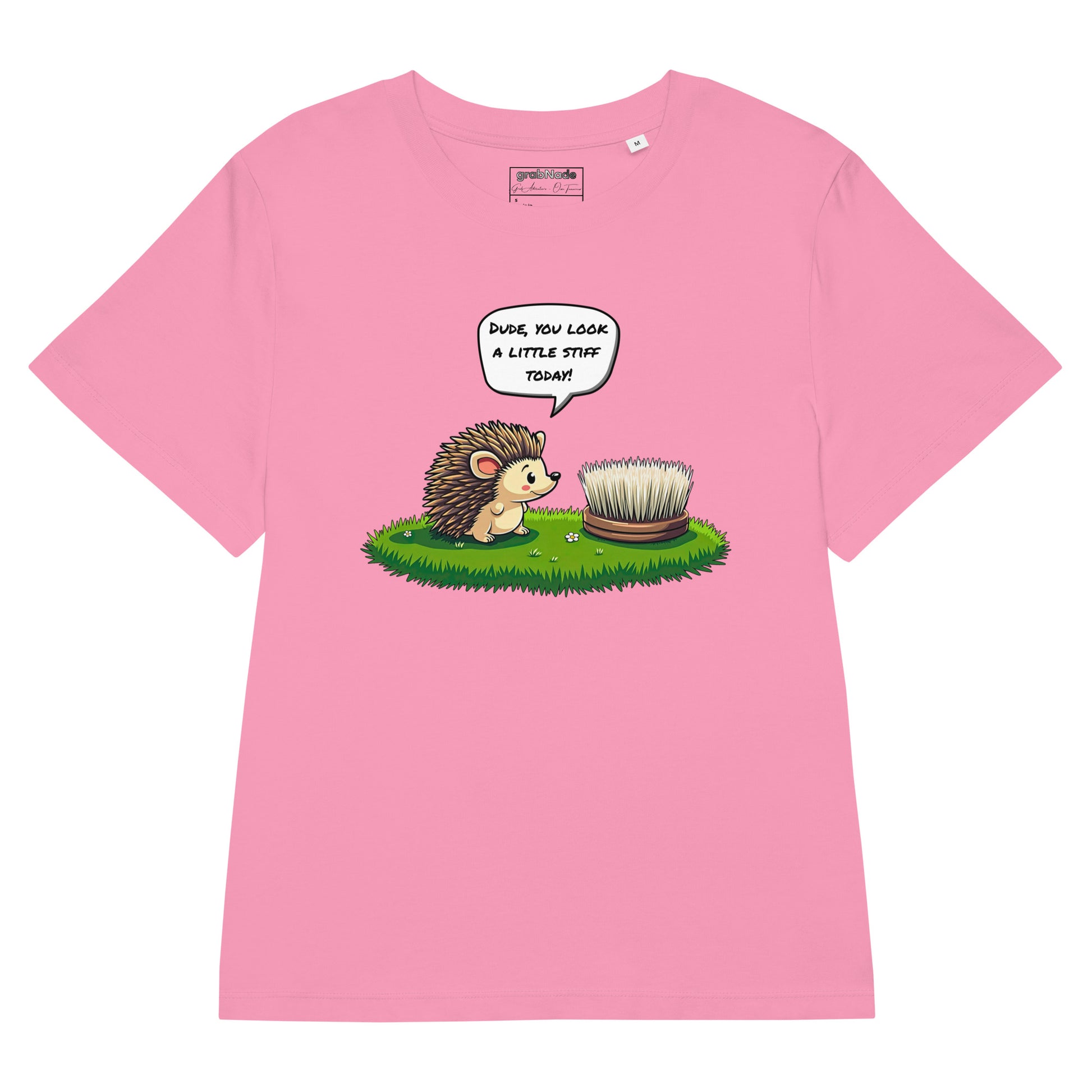 Product mockup. A flat layout pink t-shirt made from organic cotton, featuring a playful illustration on the front. The design shows a cute cartoon hedgehog standing on green grass, looking at a stiff scrubbing brush, with a speech bubble that reads, 'DUDE, YOU LOOK A LITTLE STIFF TODAY!' The 'grabNade' brand label is visible inside the neckline.