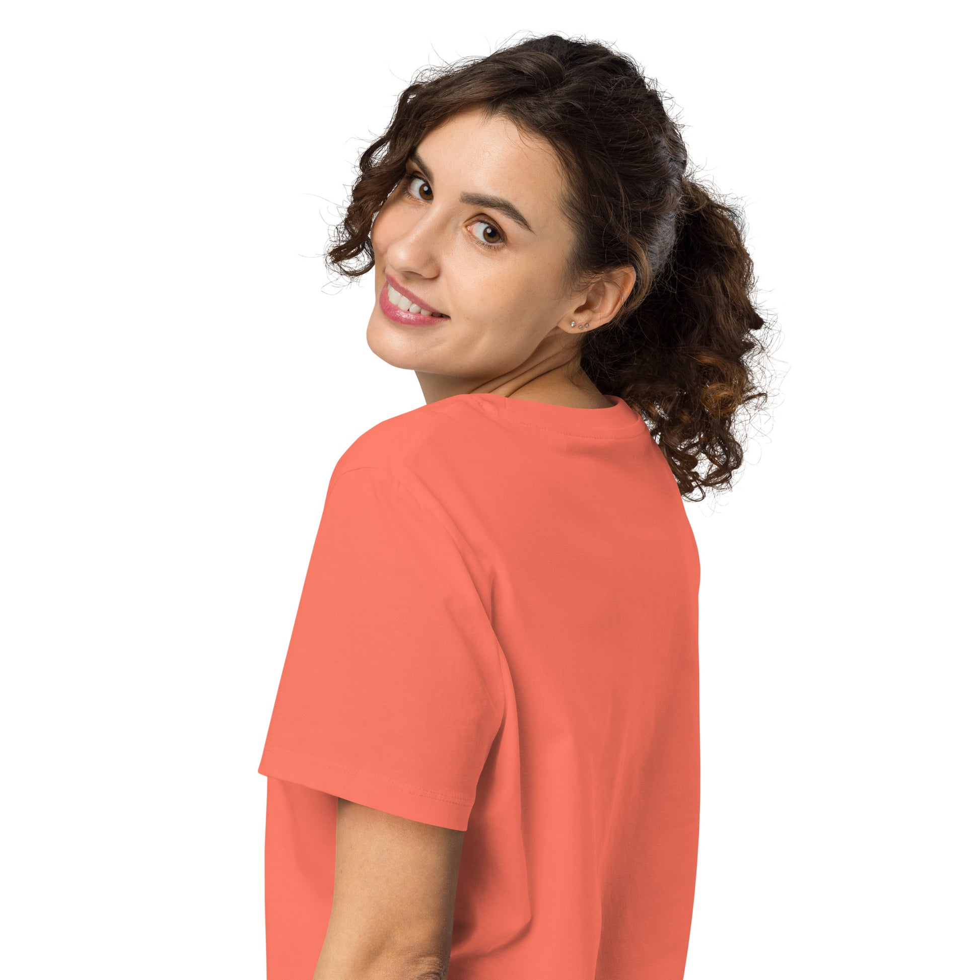 Product mockup. A woman with curly brown hair in a ponytail, shown from behind and looking over her shoulder with a smile, wearing a plain orange organic cotton t-shirt. The shirt has a soft, regular fit and no visible design on the back.
