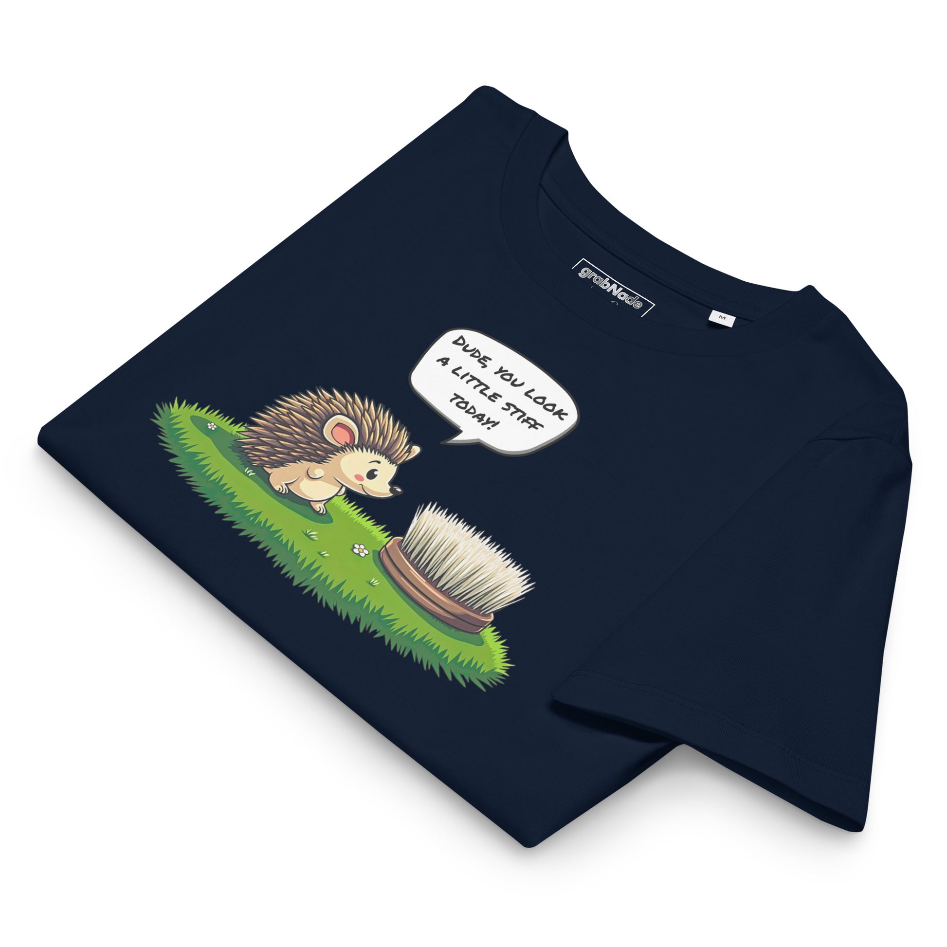 Product mockup. A folded navy blue t-shirt made from organic cotton, featuring a playful illustration on the front. The design shows a cute cartoon hedgehog standing on green grass, looking at a stiff scrubbing brush, with a speech bubble that reads, 'DUDE, YOU LOOK A LITTLE STIFF TODAY!' The 'grabNade' brand label is visible inside the neckline.