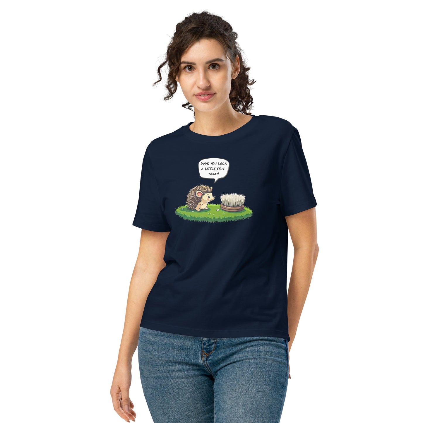 Product mockup. A happy woman wearing a navy blue organic cotton t-shirt featuring a playful cartoon illustration of a hedgehog standing on green grass beside a scrubbing brush. The hedgehog has a speech bubble that says, 'DUDE, YOU LOOK A LITTLE STIFF TODAY!'