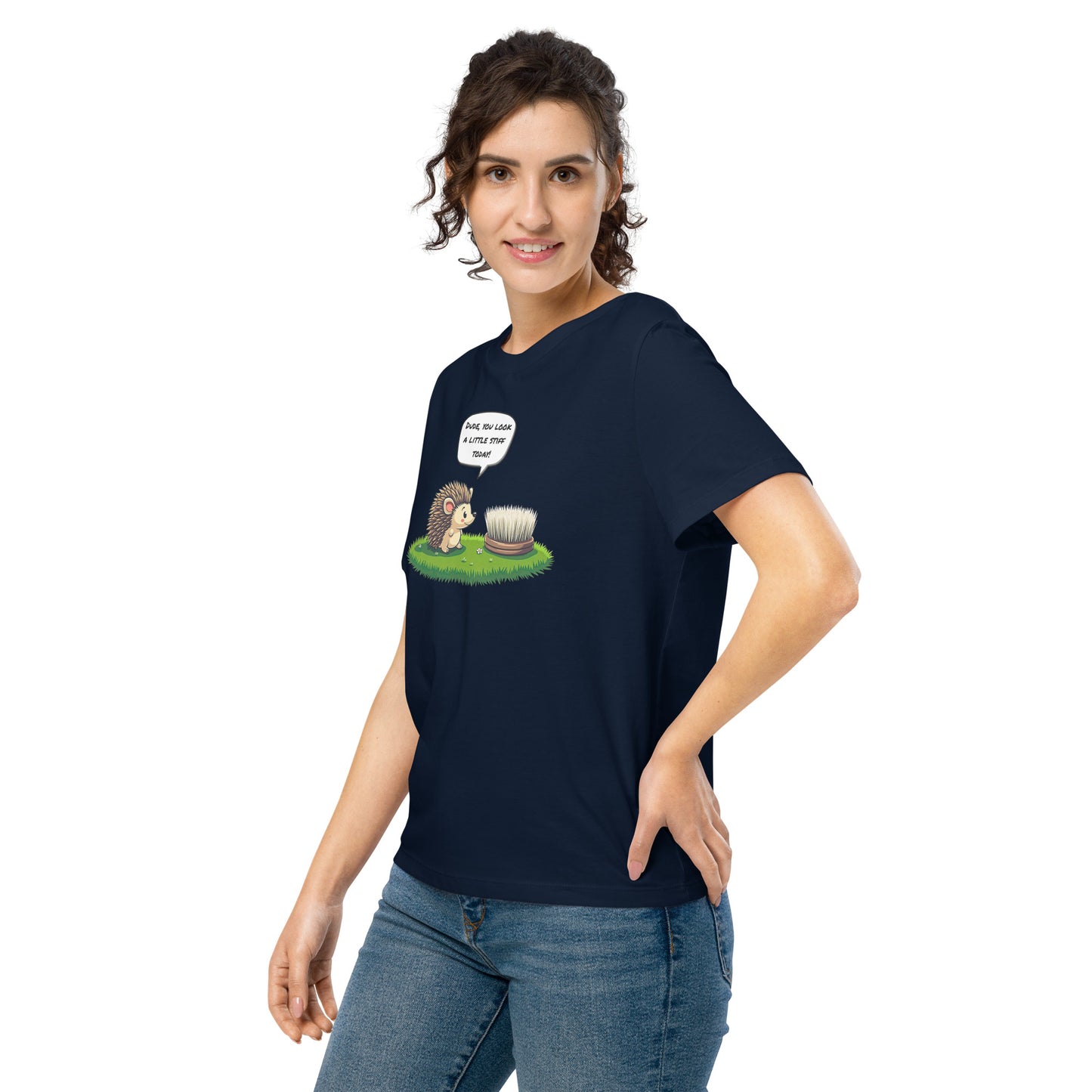 Product mockup. A happy woman wearing a navy blue organic cotton t-shirt featuring a playful cartoon illustration of a hedgehog standing on green grass beside a scrubbing brush. The hedgehog has a speech bubble that says, 'DUDE, YOU LOOK A LITTLE STIFF TODAY!'