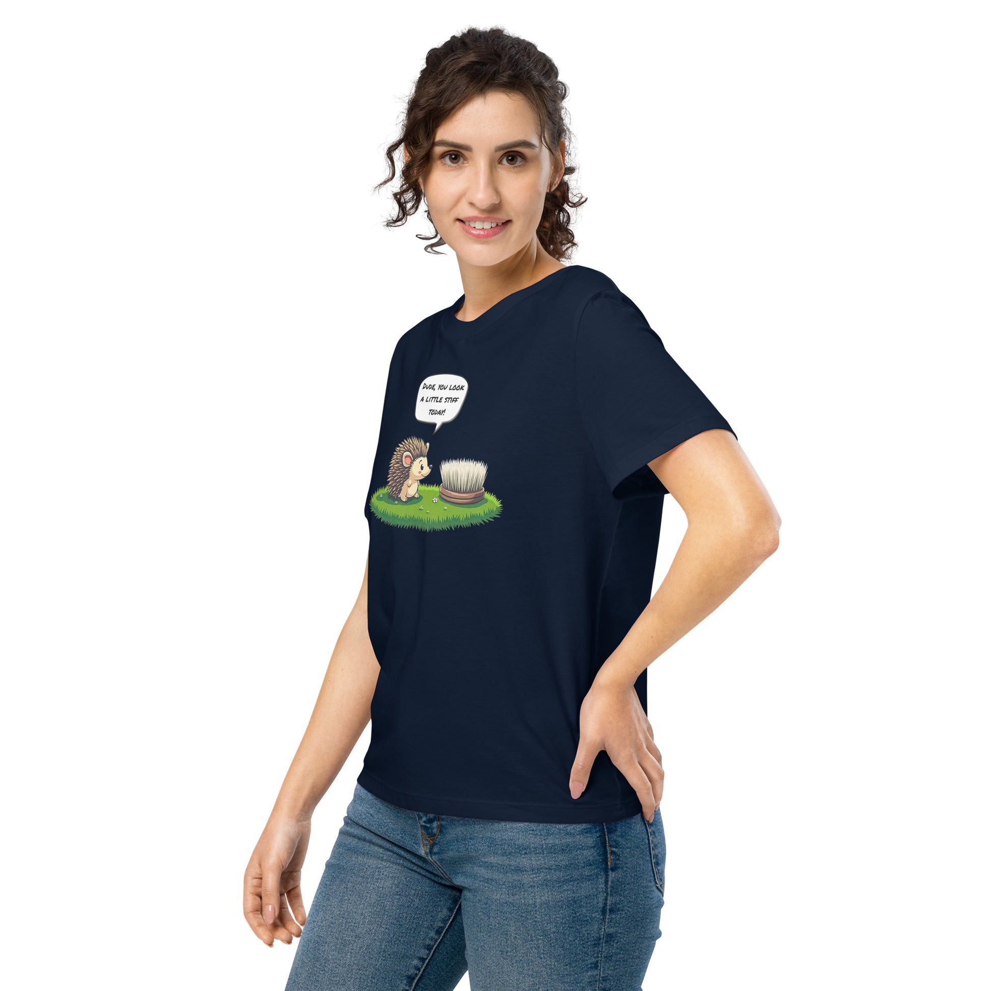 Product mockup. A happy woman wearing a navy blue organic cotton t-shirt featuring a playful cartoon illustration of a hedgehog standing on green grass beside a scrubbing brush. The hedgehog has a speech bubble that says, 'DUDE, YOU LOOK A LITTLE STIFF TODAY!'
