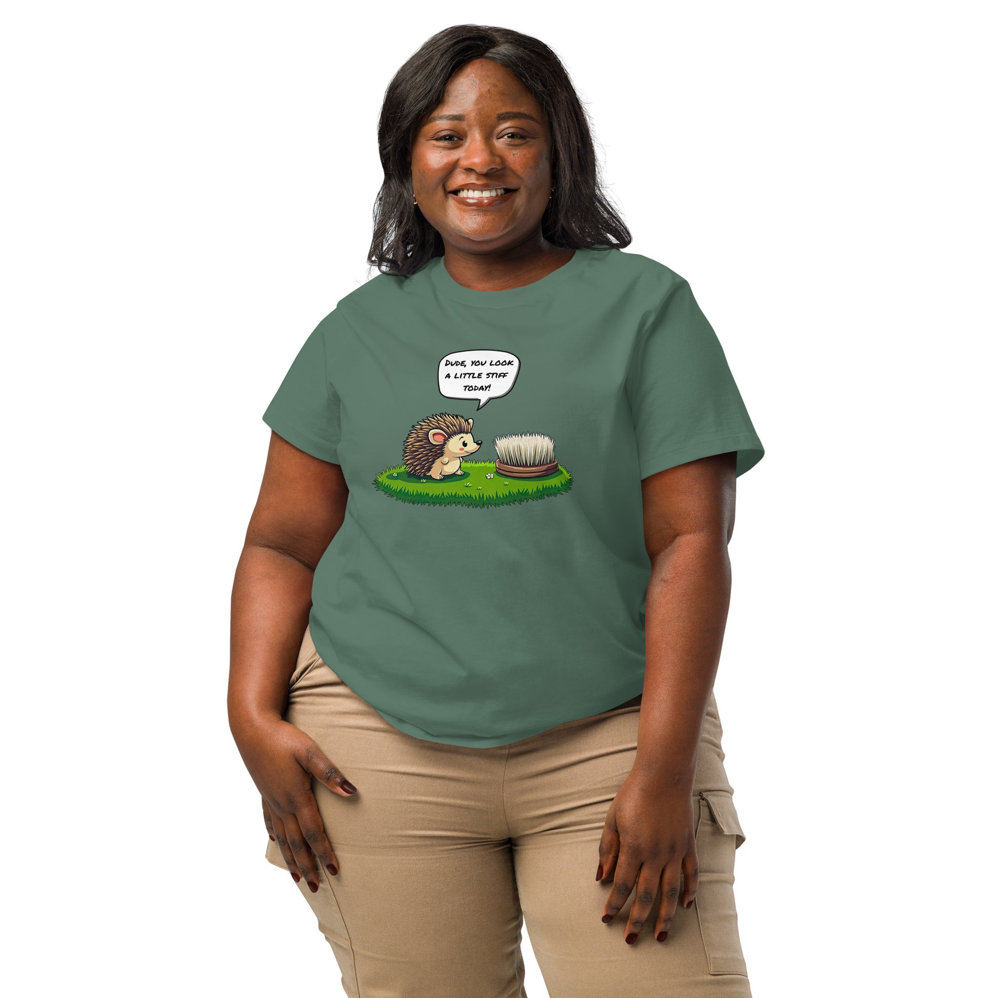 Product mockup. A happy woman wearing a 2XL green organic cotton t-shirt featuring a playful cartoon illustration of a hedgehog standing on green grass beside a scrubbing brush. The hedgehog has a speech bubble that says, 'DUDE, YOU LOOK A LITTLE STIFF TODAY!'