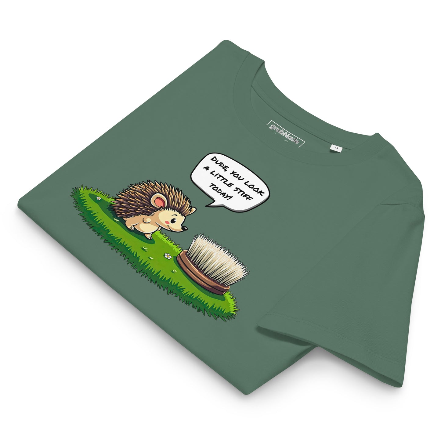 Product mockup. A folded green t-shirt made from organic cotton, featuring a playful illustration on the front. The design shows a cute cartoon hedgehog standing on green grass, looking at a stiff scrubbing brush, with a speech bubble that reads, 'DUDE, YOU LOOK A LITTLE STIFF TODAY!' The 'grabNade' brand label is visible inside the neckline.