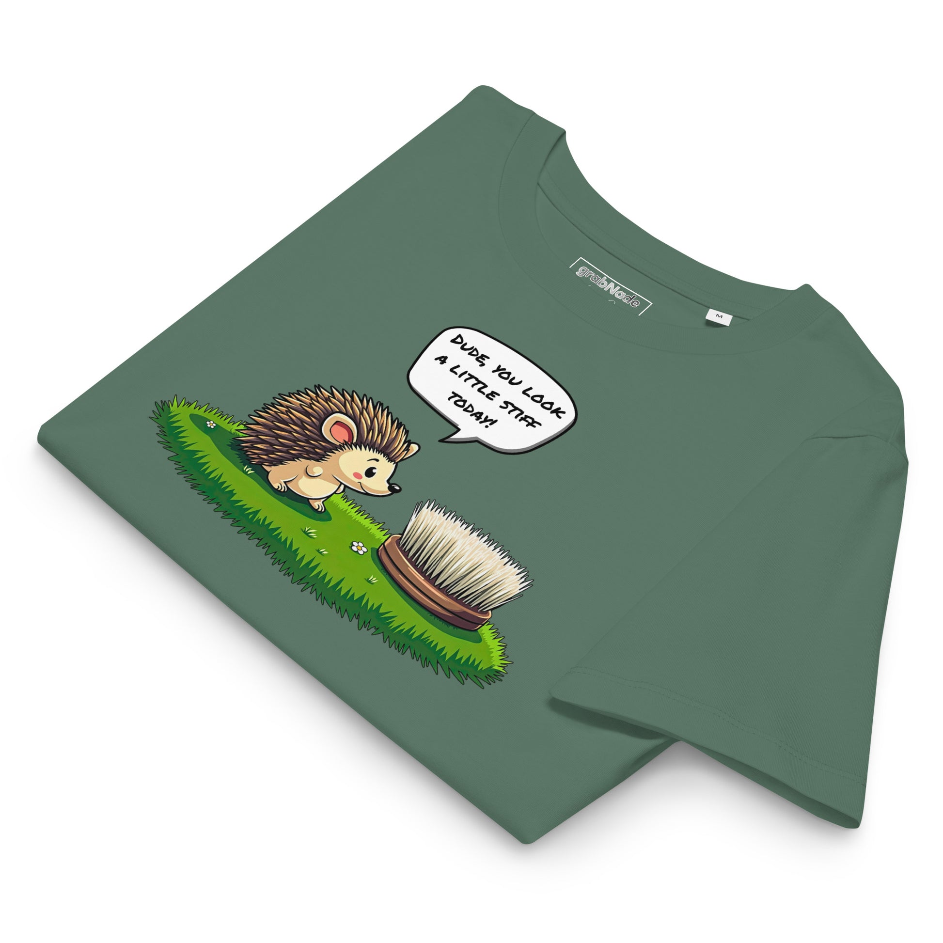 Product mockup. A folded green t-shirt made from organic cotton, featuring a playful illustration on the front. The design shows a cute cartoon hedgehog standing on green grass, looking at a stiff scrubbing brush, with a speech bubble that reads, 'DUDE, YOU LOOK A LITTLE STIFF TODAY!' The 'grabNade' brand label is visible inside the neckline.