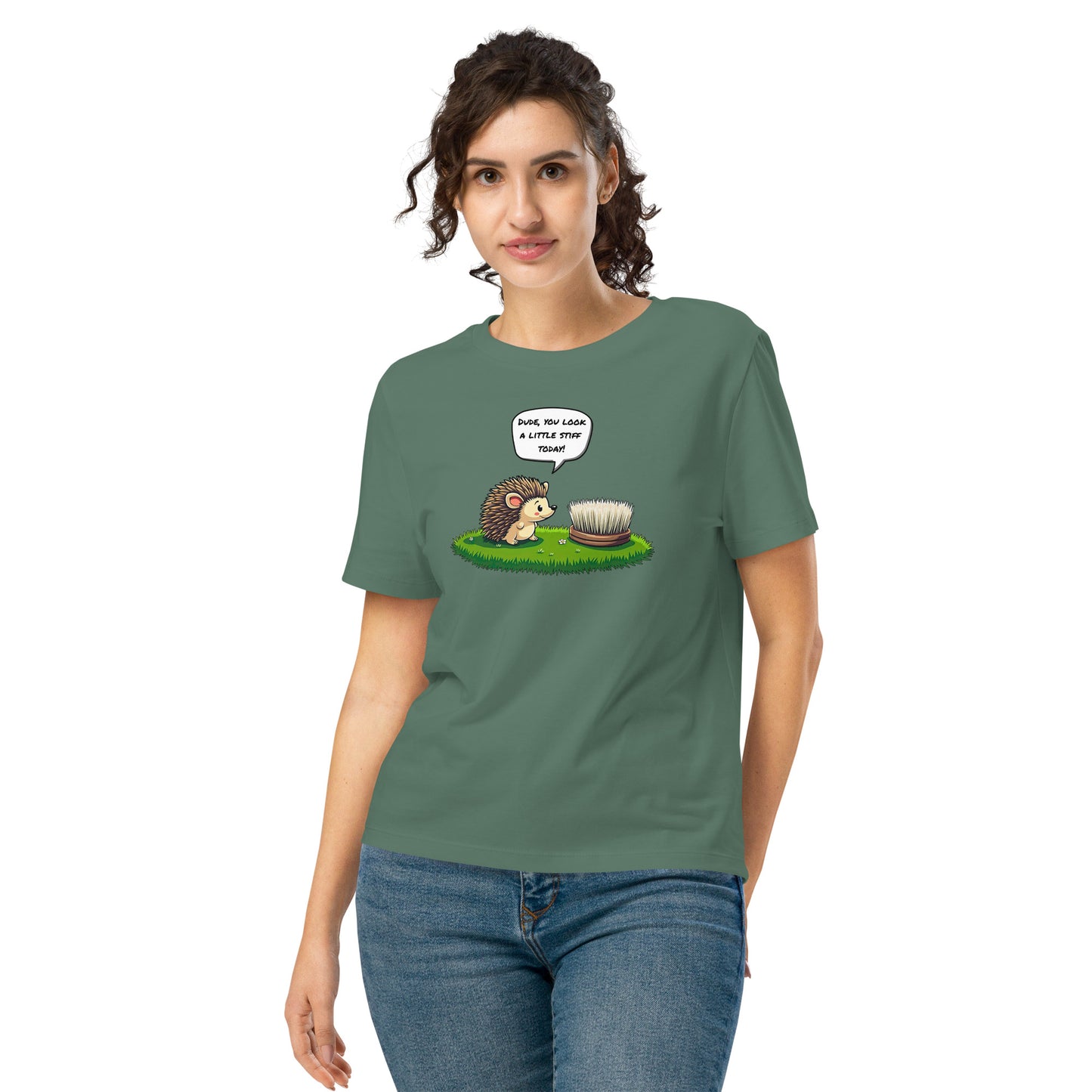 Product mockup. A happy woman wearing a green organic cotton t-shirt featuring a playful cartoon illustration of a hedgehog standing on green grass beside a scrubbing brush. The hedgehog has a speech bubble that says, 'DUDE, YOU LOOK A LITTLE STIFF TODAY!'